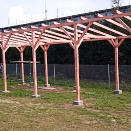 Surowa konstrukcja drewnianej altany z widocznymi podporami i belkami, ustawiona na betonowych fundamentach, z zielonymi rusztowaniami w tle, na tle metalowej siatki ogrodzeniowej i drzew