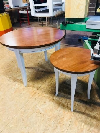 Dwa owalne stoliki z drewnianymi blatami i białymi nogami, większy i mniejszy, widoczne w warsztacie na tle maszyn stolarskich i elementów mebli.