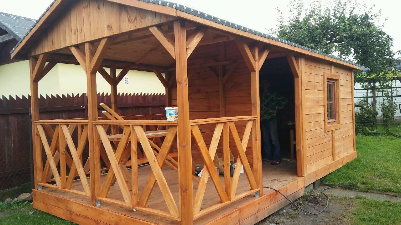 Drewniana altana z balustradą w kształcie litery X, widoczny fragment dachu, okno i osoba wewnątrz, na trawniku