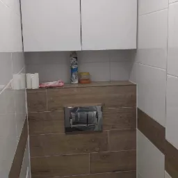 Nowoczesna toaleta podwieszana z zabudową, białe płytki z brązowym pasem, drewnopodobna obudowa stelaża, białe szafki wiszące nad toaletą, widoczne środki czystości i szczotka do WC.