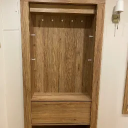 Wnęka z drewnianą zabudową: szafka z szufladą, półka na ubrania z wieszakami, wykończona w fornirze o ciepłym odcieniu drewna, z oświetleniem ściennym w tle.