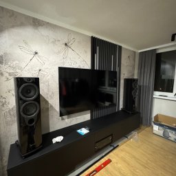 DiPol Meble - Dmytro Polishchuk - Czarna szafka RTV z kolumnami głośnikowymi na tle ściany z motywem ważek. Widoczne narzędzia sugerują montaż lub prace wykończeniowe.