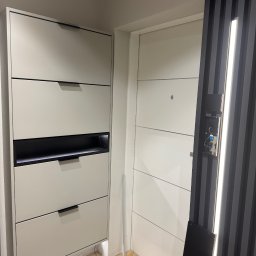 DiPol Meble - Dmytro Polishchuk - Jasnoszara szafka na buty z czterema szufladami i czarnymi uchwytami, obok białych drzwi z zamkiem elektronicznym i czarną, żebrowaną ścianką działową.