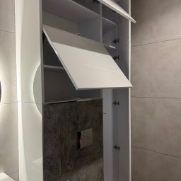 DiPol Meble - Dmytro Polishchuk - Biała szafka łazienkowa z otwartymi drzwiczkami, widoczne półki wewnątrz. Na ścianie szare płytki z dekoracyjnym wzorem. Nowoczesna aranżacja wnętrza łazienki.