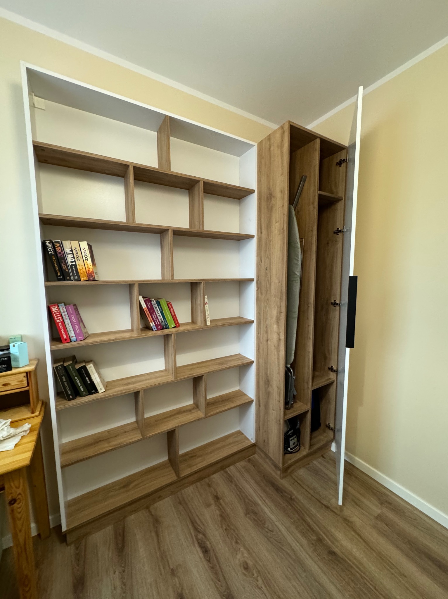 Wnętrze pokoju z zabudową meblową: regał z drewnianymi półkami i białą ramą, częściowo wypełniony książkami, po prawej szafa z otwartymi drzwiami, w której widoczny jest odkurzacz i deska...