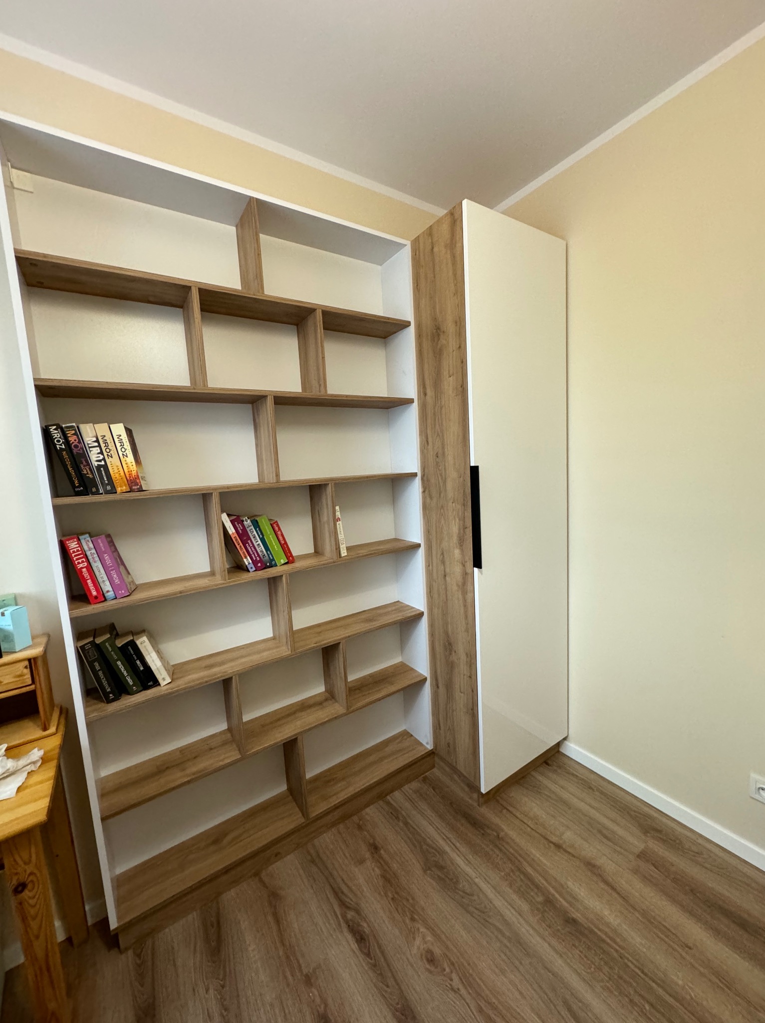 Biało-drewniana zabudowa meblowa z półkami, niektóre z książkami, połączona z wysoką szafką z czarnym uchwytem, na tle beżowej ściany i drewnopodobnej podłogi.