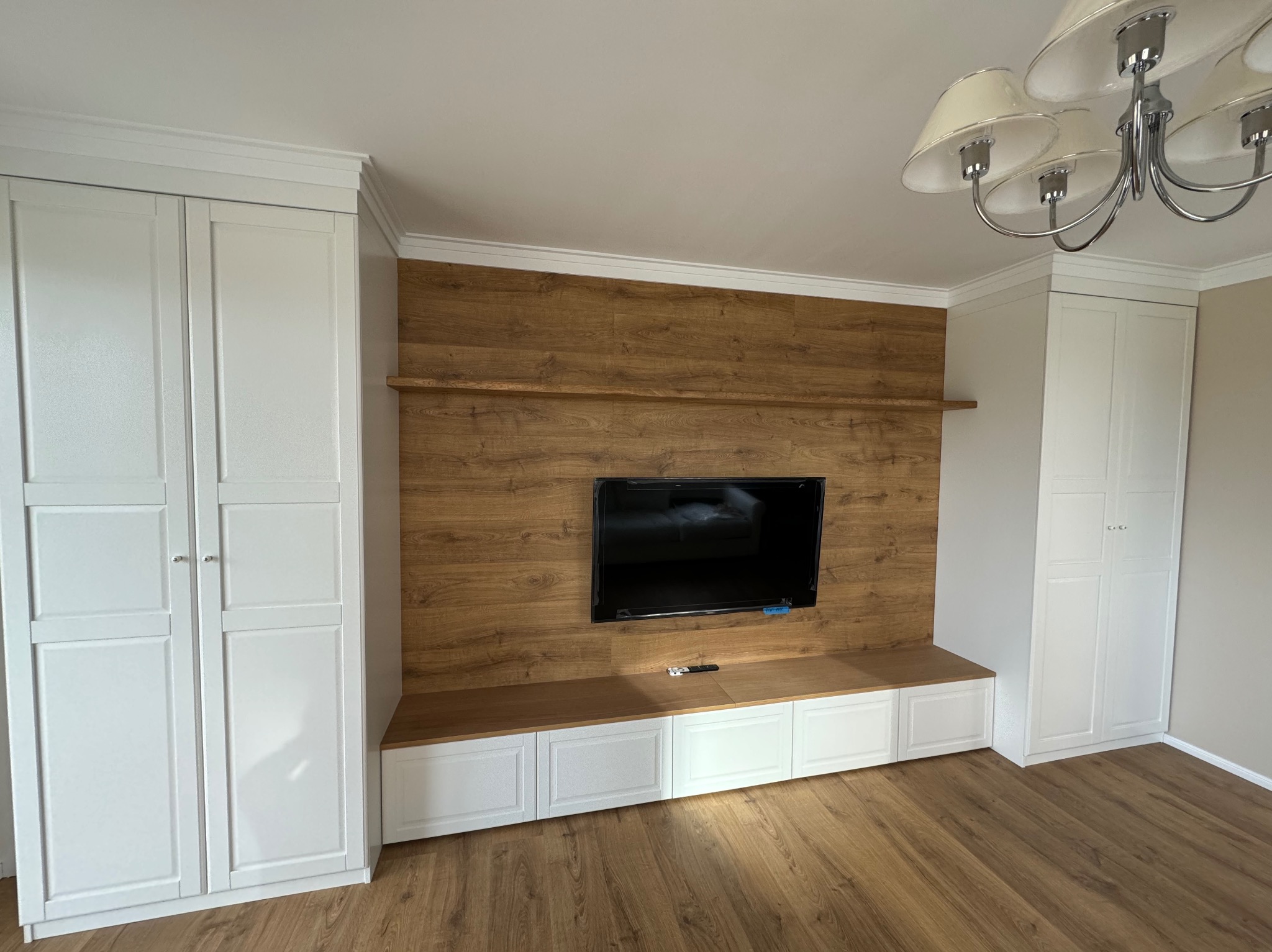 Białe szafy wnękowe z drewnianym panelem ściennym i szafką RTV pod telewizorem, oświetlone przez klasyczny żyrandol.