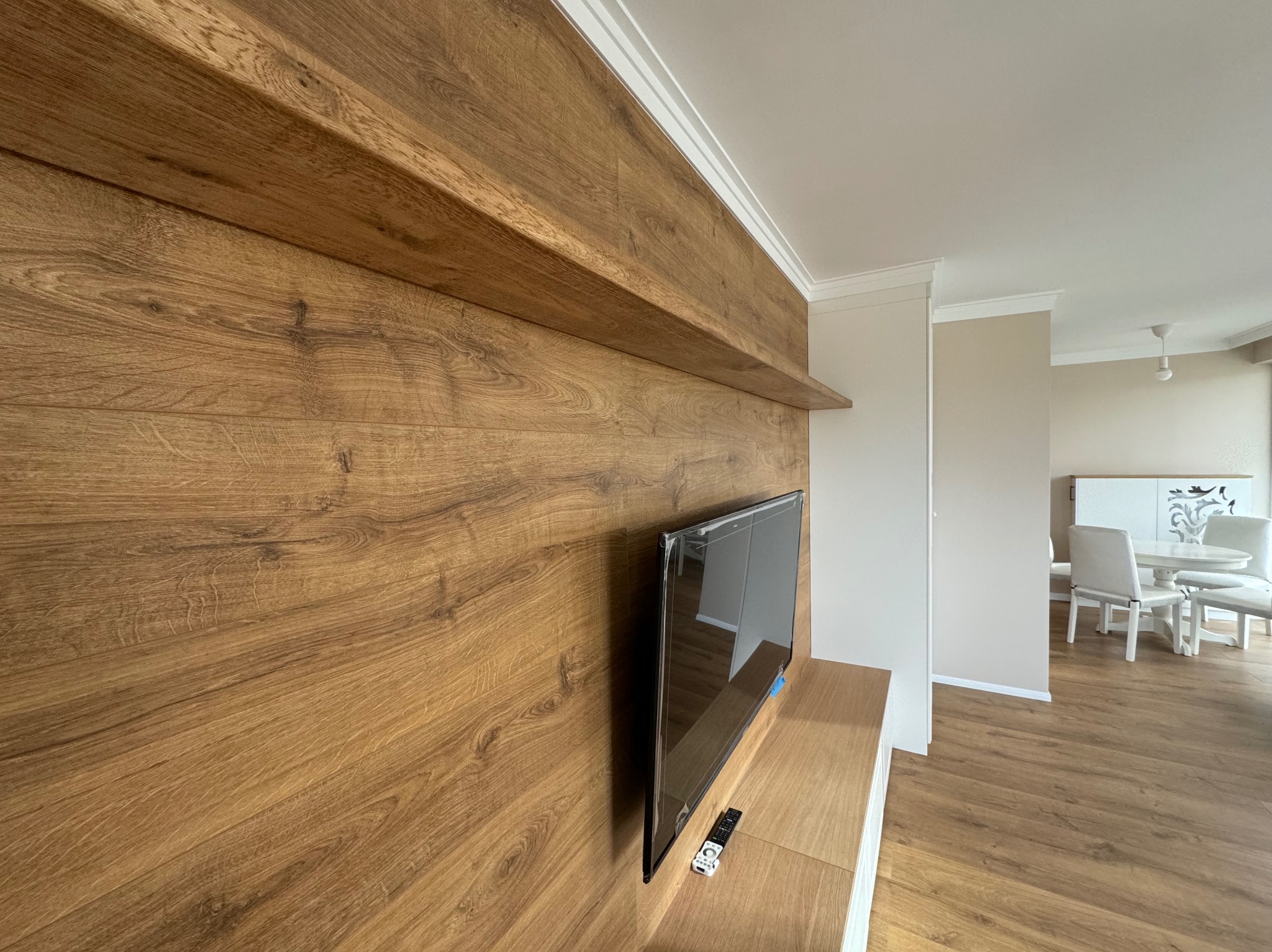 Drewniana ściana z półkami i zamontowanym telewizorem w salonie z widoczną jadalnią w tle.