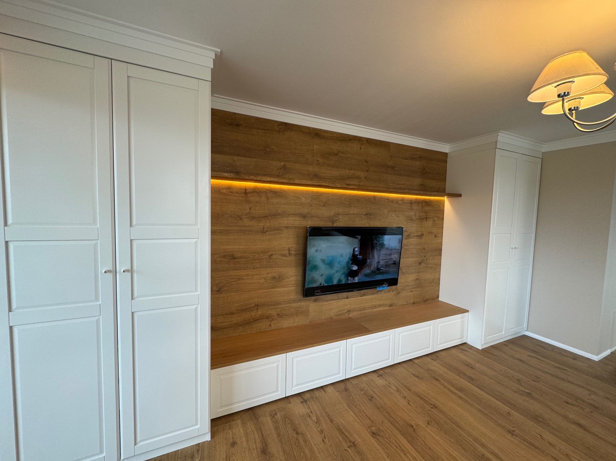 Salon z zabudową meblową: białe szafy z frezowanymi frontami po bokach, centralna ściana z drewnopodobną okładziną, podświetleniem LED i telewizorem, niska szafka RTV z blatem z drewna.