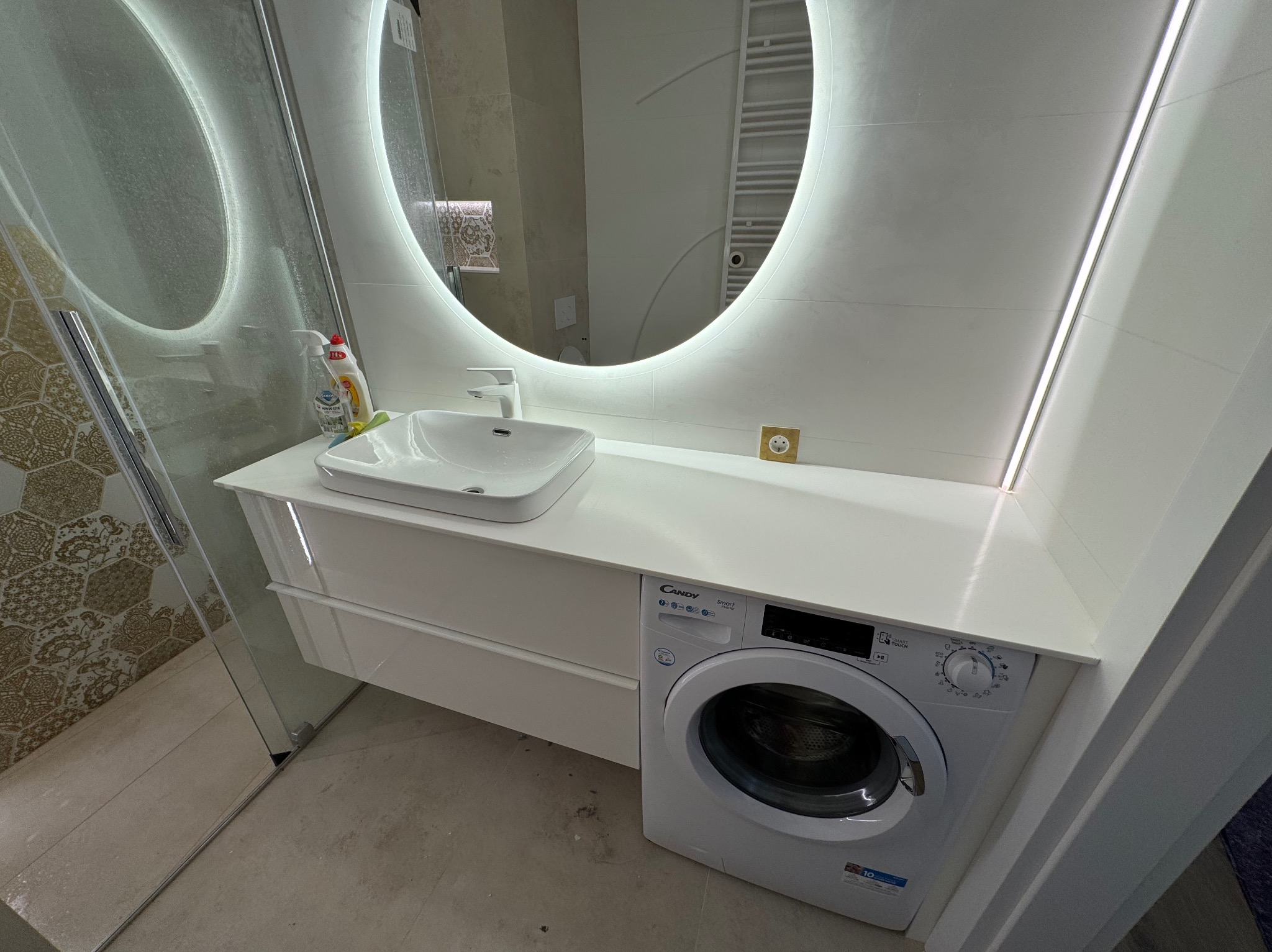 Nowoczesna łazienka z umywalką nablatową na białej szafce, podświetlanym okrągłym lustrem i pralką Candy Smart Touch w zabudowie.
