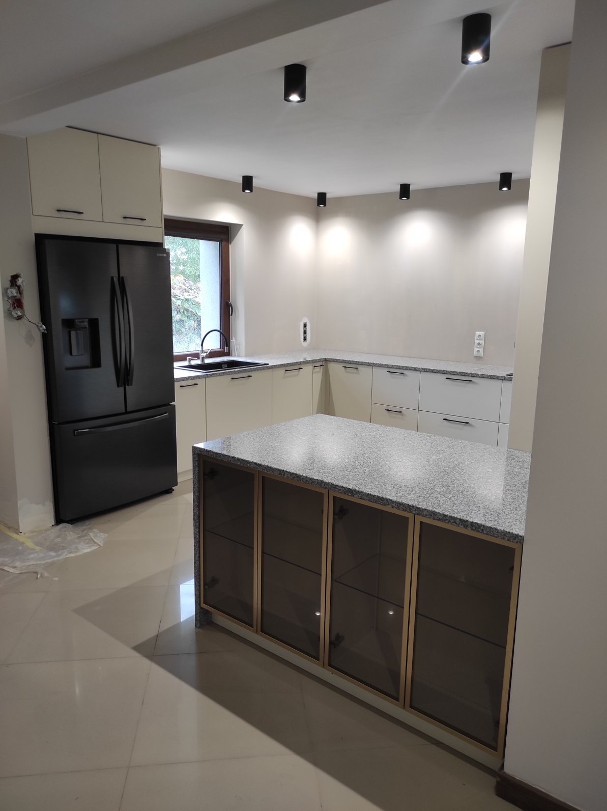 Nowoczesna kuchnia z wyspą i czarną lodówką w Oławie. Beżowe szafki z ciemnymi uchwytami, szary blat i punktowe oświetlenie sufitowe.