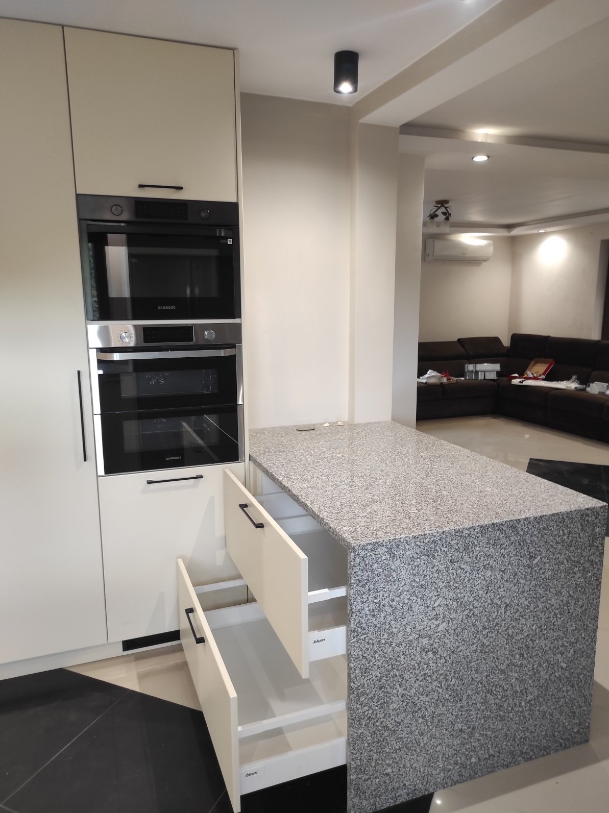Nowoczesna kuchnia z kremowymi frontami, zabudowanymi piekarnikami Samsung i wyspą z szarym, nakrapianym blatem. Dwie szuflady wyspy są otwarte, ukazując system Blum.