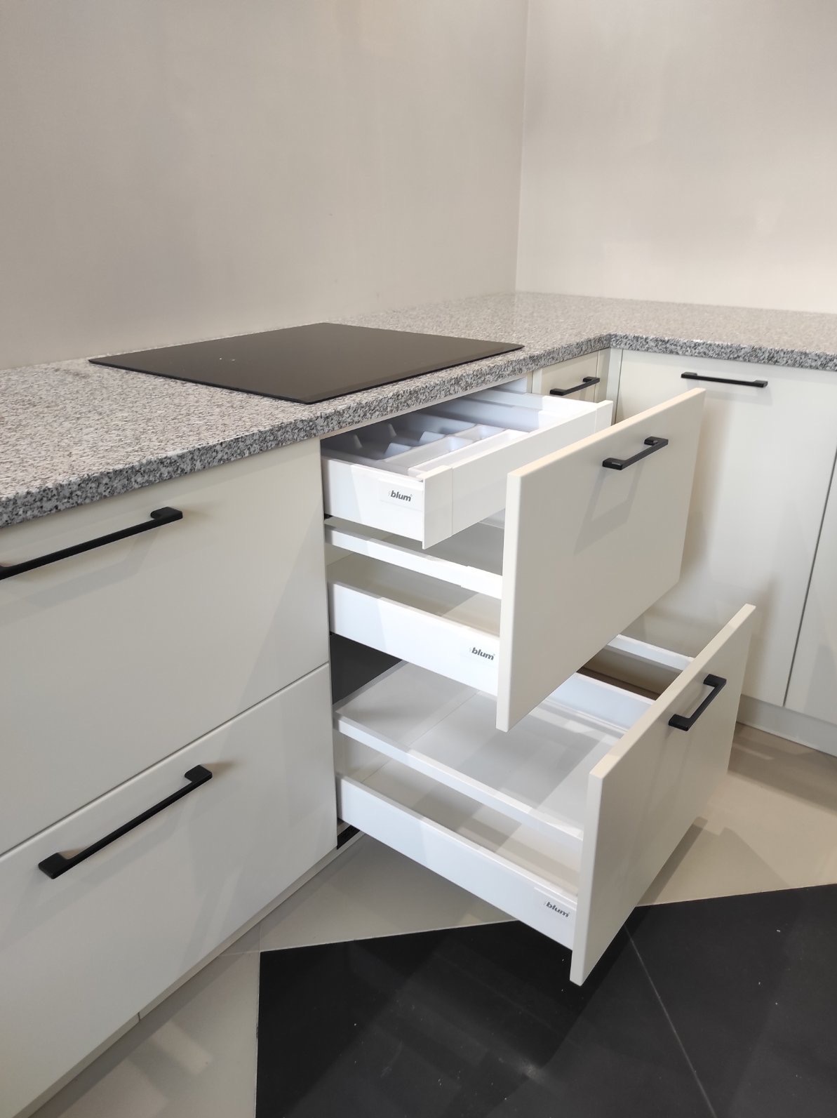 Białe szuflady kuchenne z czarnymi uchwytami, z widocznym systemem organizacji sztućców w górnej szufladzie, blat z szarego granitu, płyta indukcyjna.