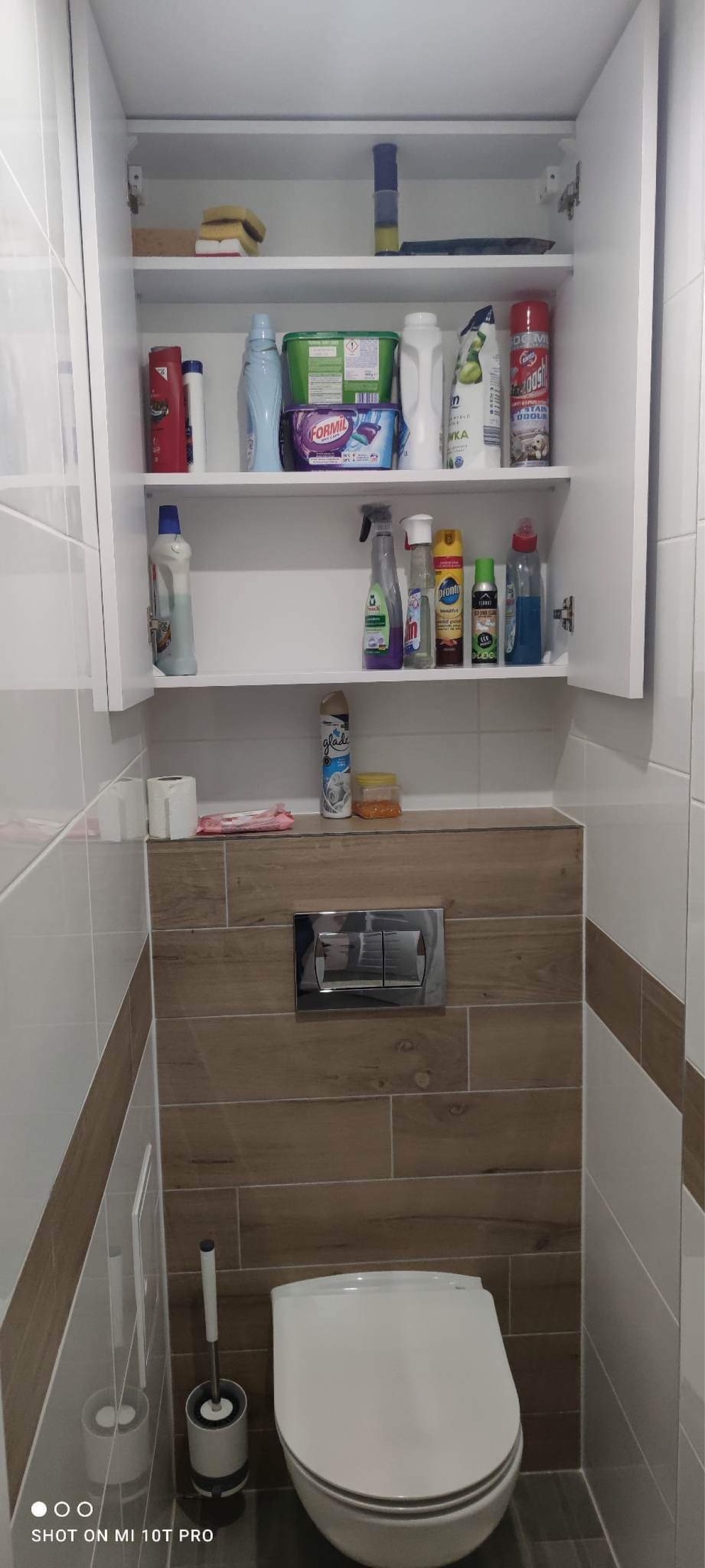 Biała szafka łazienkowa nad toaletą, wypełniona środkami czystości i akcesoriami, z drewnopodobną obudową i chromowanym przyciskiem spłuczki.