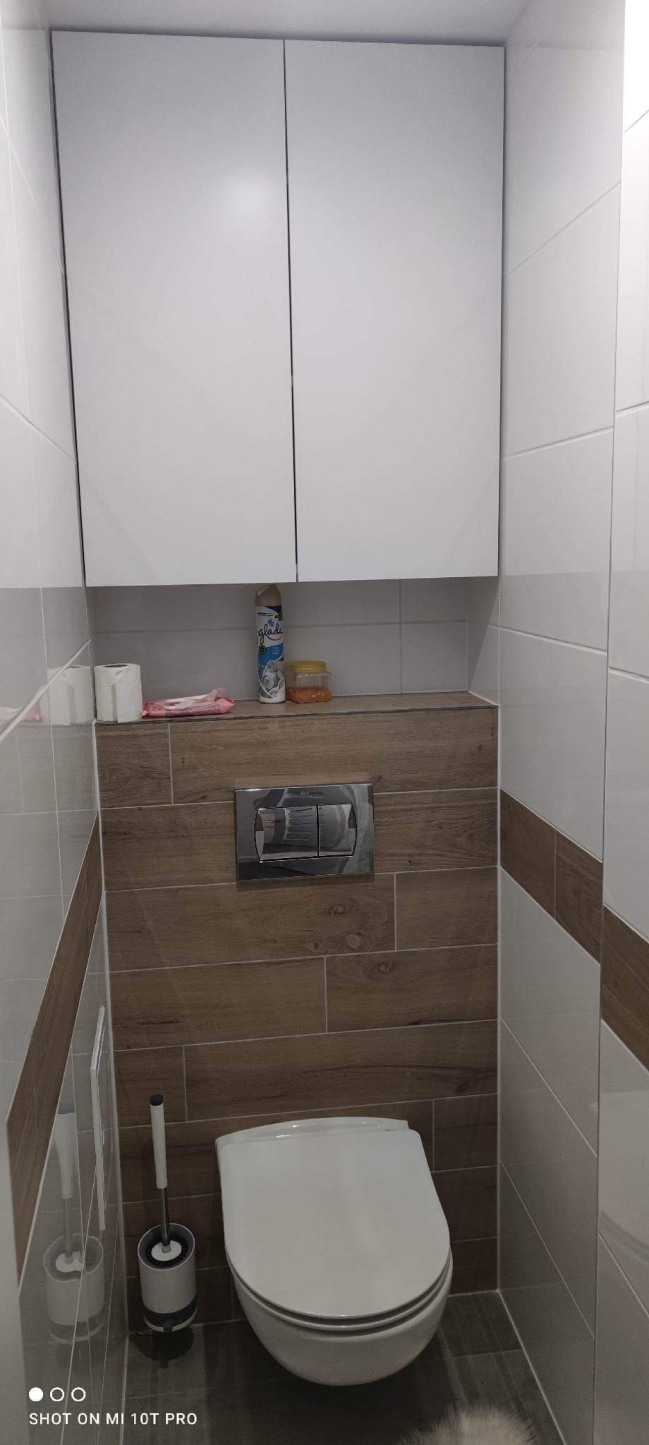 Nowoczesna toaleta podwieszana z zabudową, białe płytki z brązowym pasem, drewnopodobna obudowa stelaża, białe szafki wiszące nad toaletą, widoczne środki czystości i szczotka do WC.