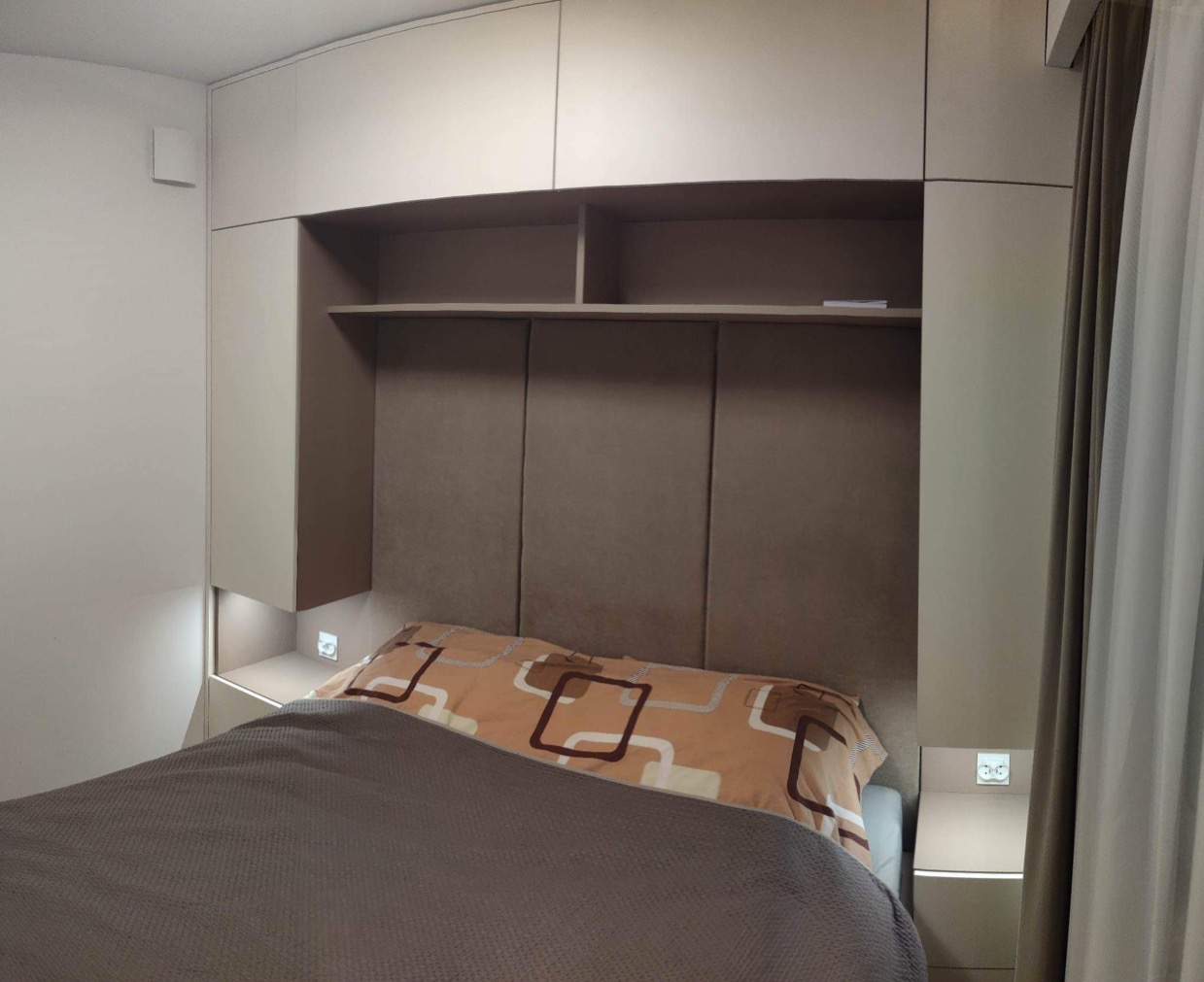 Beżowa zabudowa meblowa nad łóżkiem z półkami i oświetleniem w sypialni w Oławie.