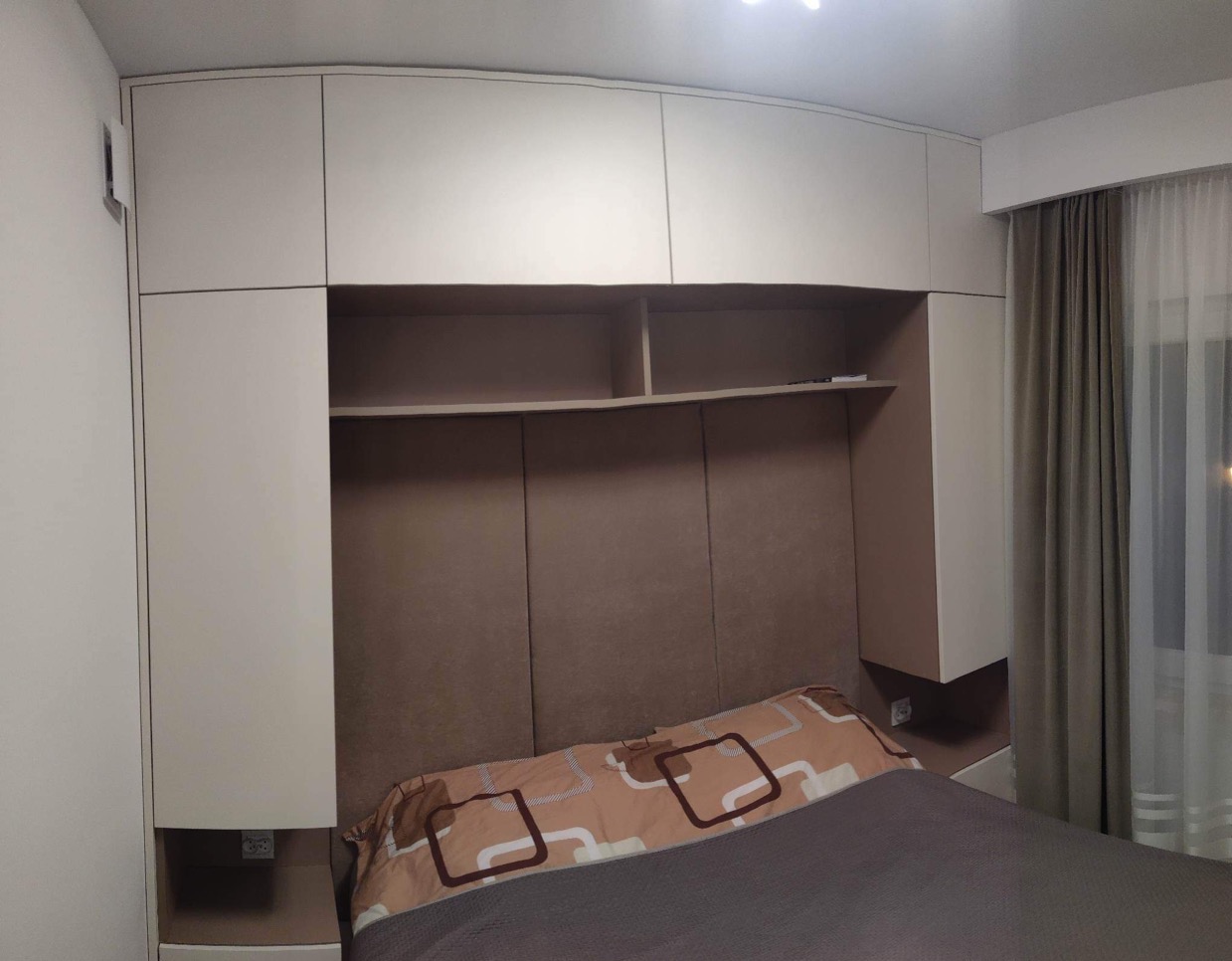 Jasna zabudowa meblowa nad łóżkiem z szafkami górnymi, półkami i panelami tapicerowanymi w kolorze beżowym, z widocznym fragmentem zasłony.