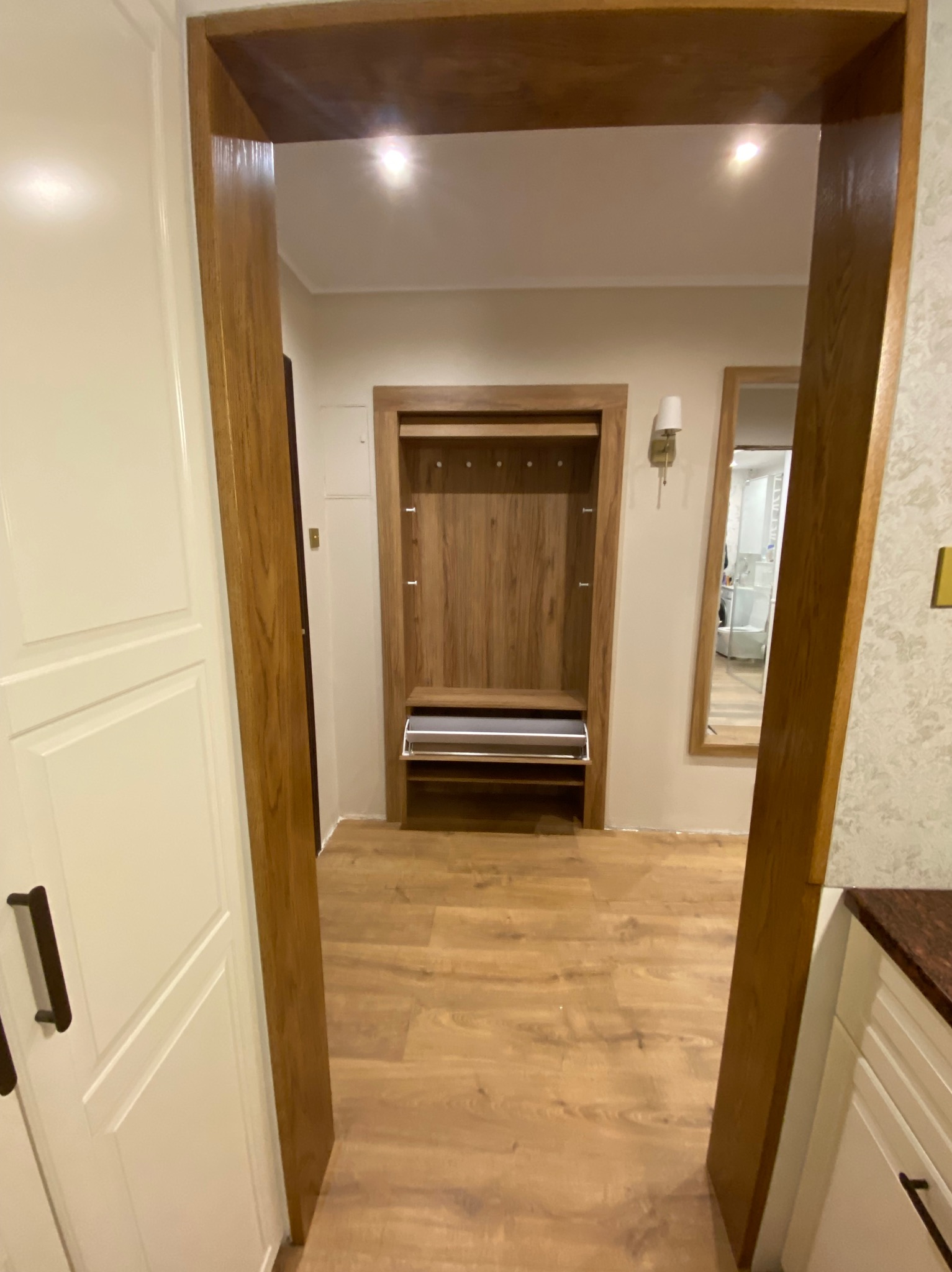 Widok z perspektywy wejścia na zabudowaną garderobę z drewnianymi elementami, w tle lustro i lampa ścienna.