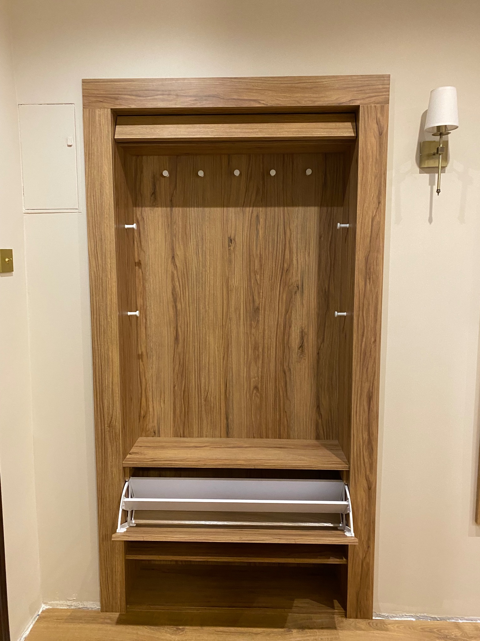 Wnęka w przedpokoju z drewnianą obudową, wyposażona w wieszaki na ubrania i wysuwany kosz na buty.