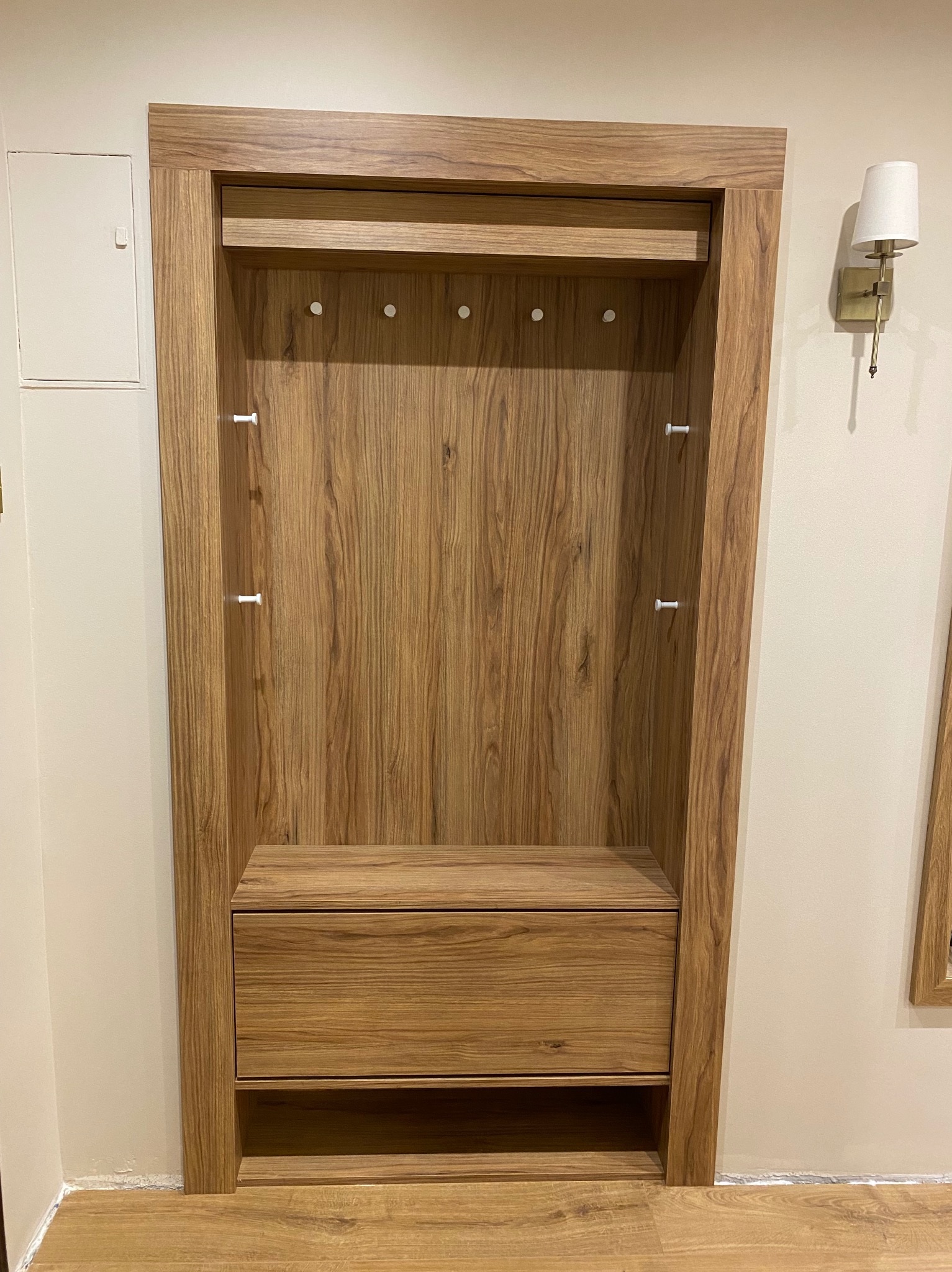 Wnęka z drewnianą zabudową: szafka z szufladą, półka na ubrania z wieszakami, wykończona w fornirze o ciepłym odcieniu drewna, z oświetleniem ściennym w tle.