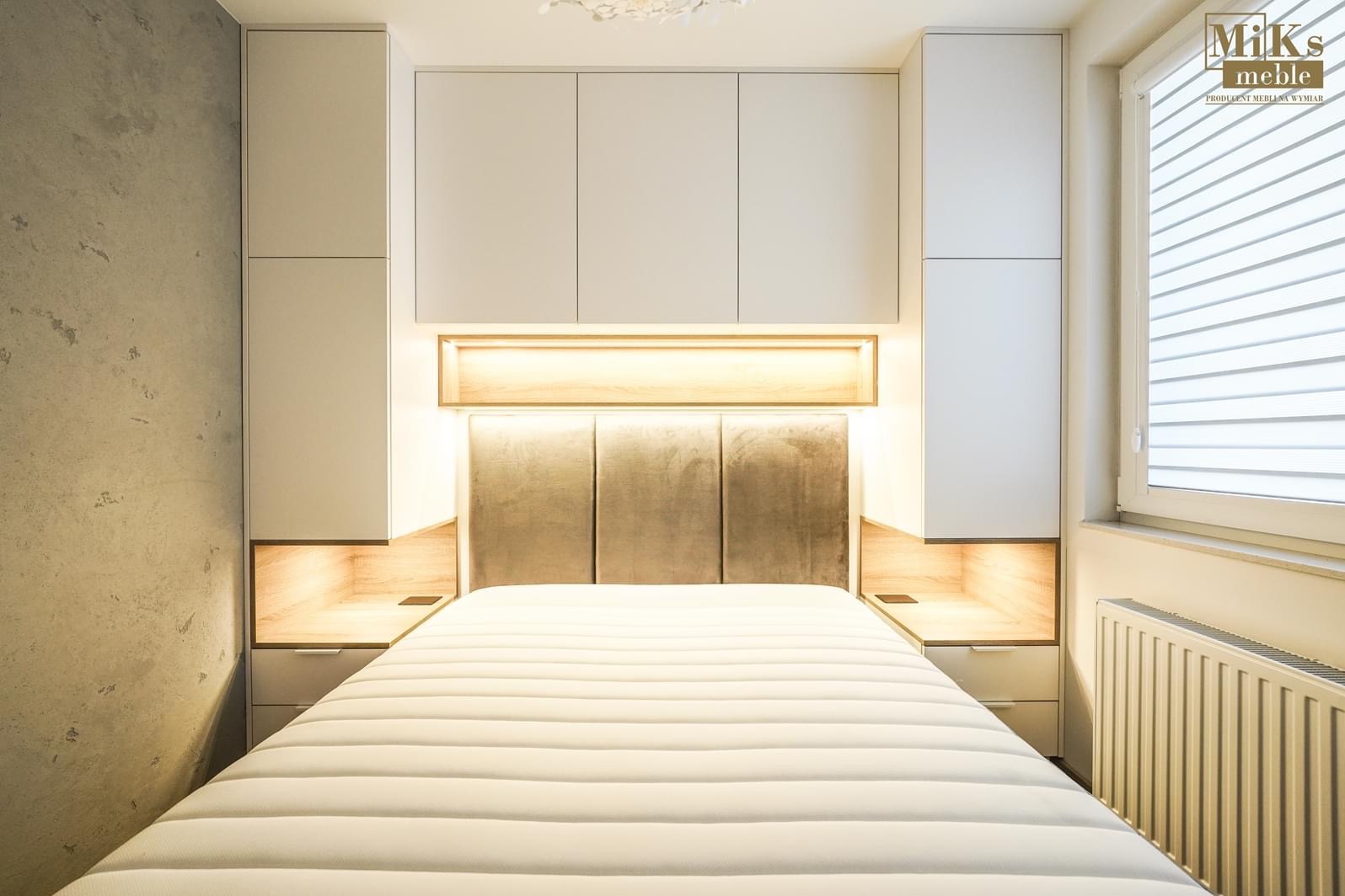 Nowoczesna sypialnia z zabudową meblową nad łóżkiem, obejmującą szafki, półki z oświetleniem LED i tapicerowany zagłówek w odcieniach szarości, uzupełniona o szafki nocne z drewnianymi akcentami.