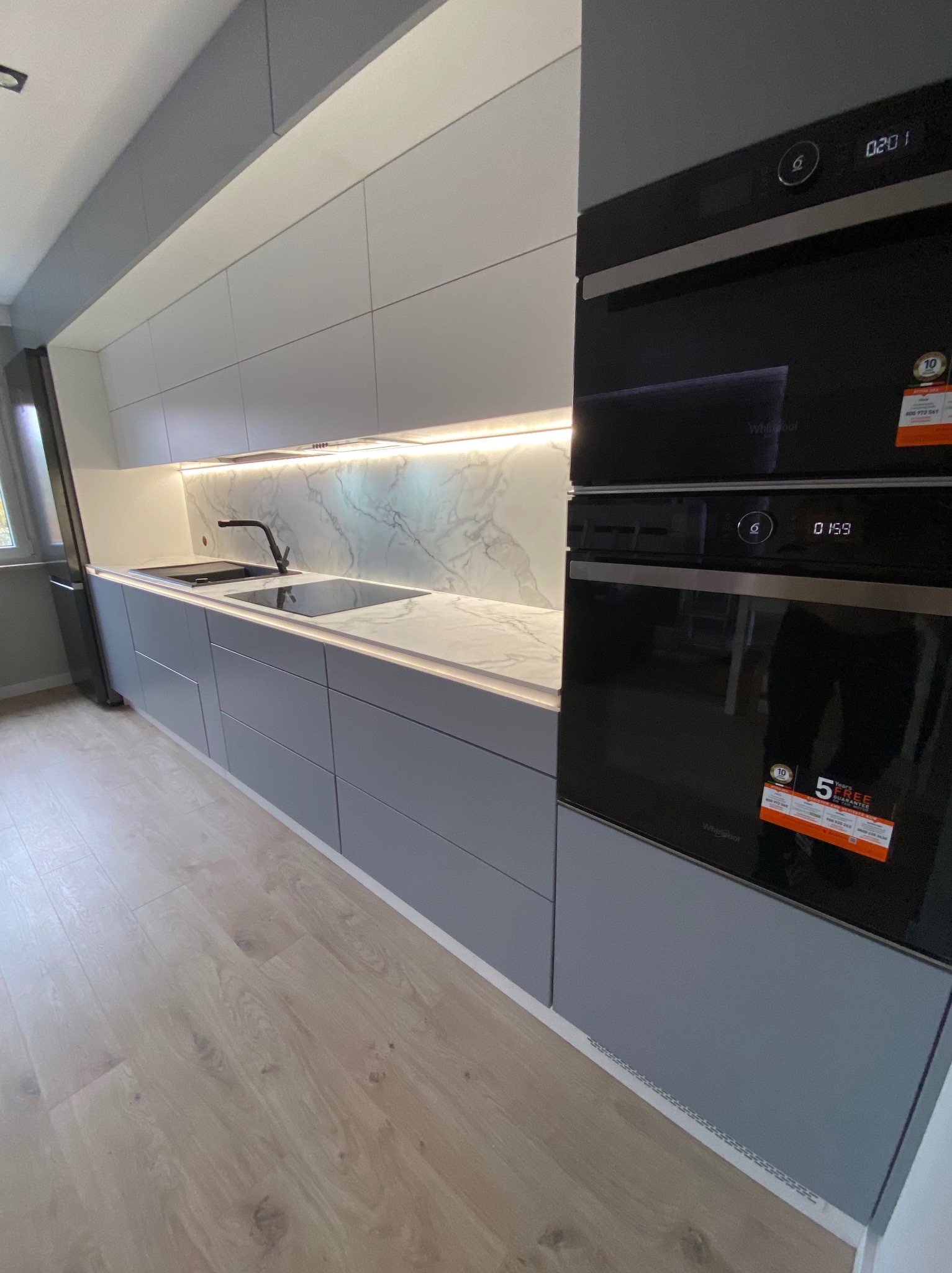Nowoczesna kuchnia z szarymi szafkami, blatem imitującym marmur, podświetleniem LED i wbudowanym piekarnikiem Whirlpool z widocznymi naklejkami gwarancyjnymi.