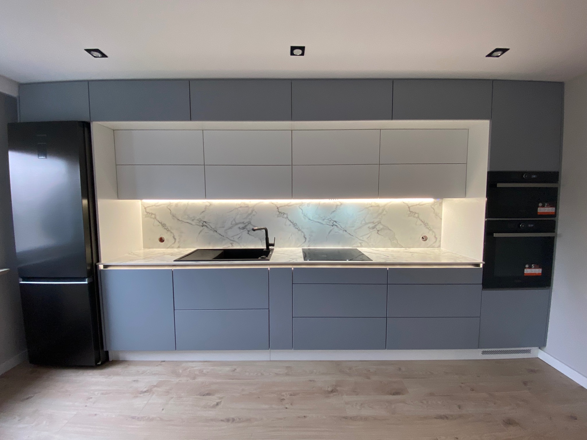 Nowoczesna kuchnia z szarymi szafkami, białymi górnymi modułami i marmurowym blatem, oświetlona listwą LED, z czarną lodówką po lewej stronie i wbudowanym piekarnikiem po prawej.