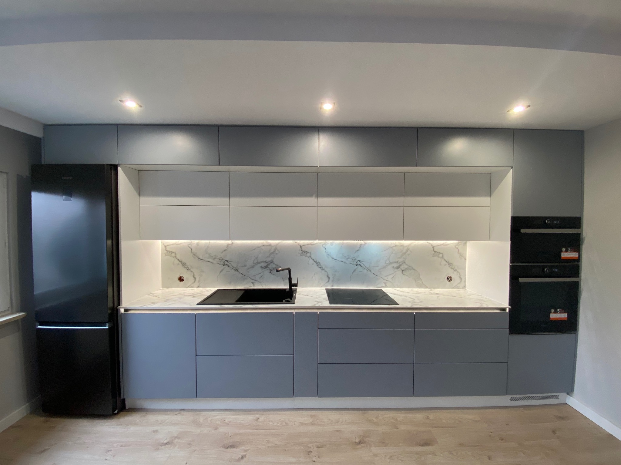 Nowoczesna kuchnia w odcieniach szarości z białym marmurowym backsplashiem, czarnym zlewem i płytą indukcyjną, oświetleniem podszafkowym LED i zabudowaną lodówką.