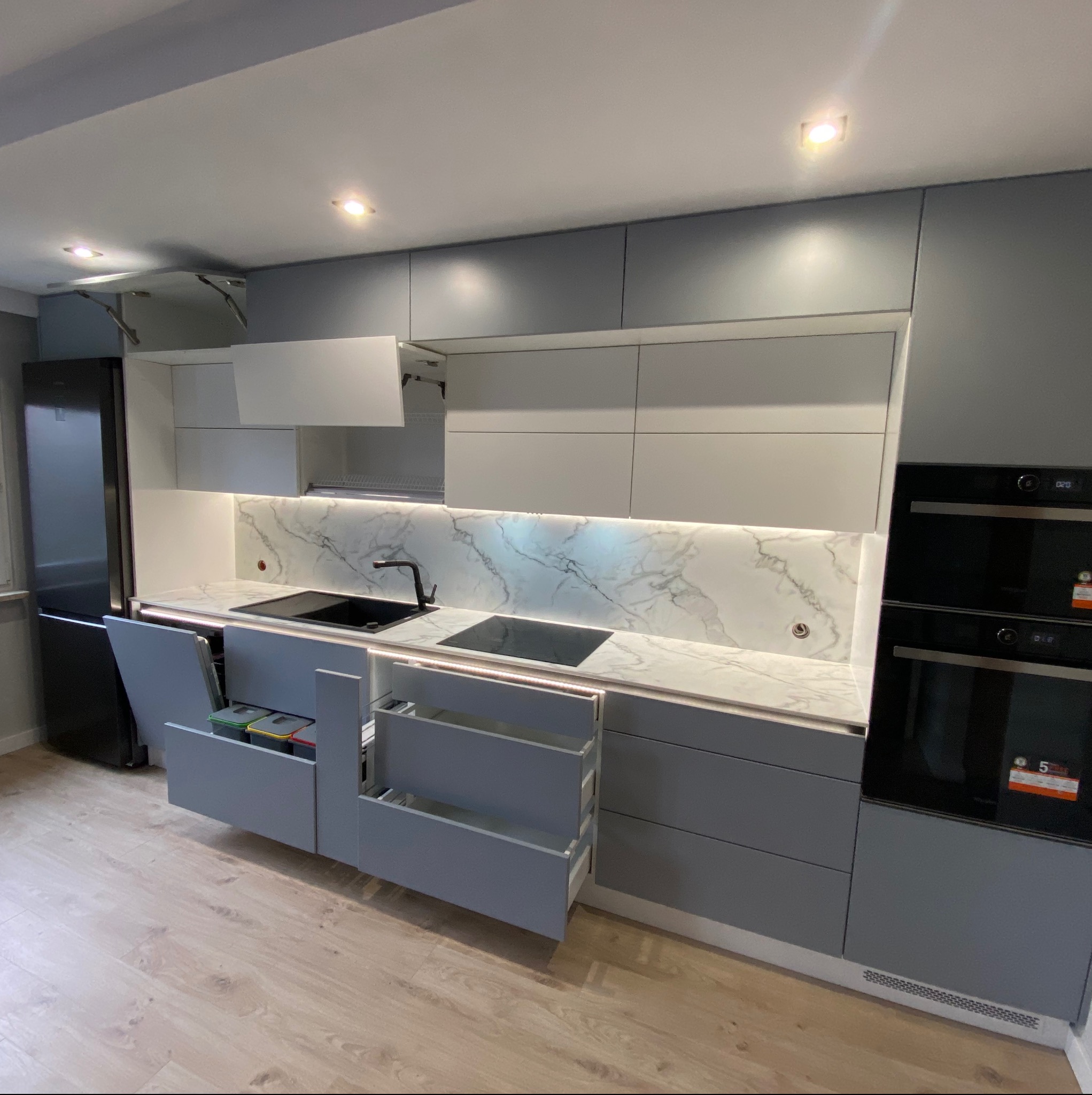 Nowoczesna, szara kuchnia z marmurowym panelem nad blatem, otwartymi szufladami z systemem sortowania odpadów i szufladami cargo, oświetleniem LED pod szafkami i w szufladach, oraz wbudowanym...