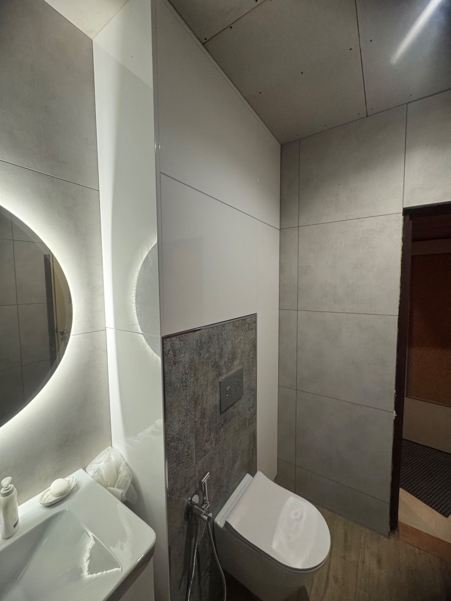 Nowoczesna łazienka z podświetlanym lustrem, szarą glazurą i białymi szafkami w zabudowie. Widoczna toaleta i umywalka z armaturą. Minimalistyczny design.