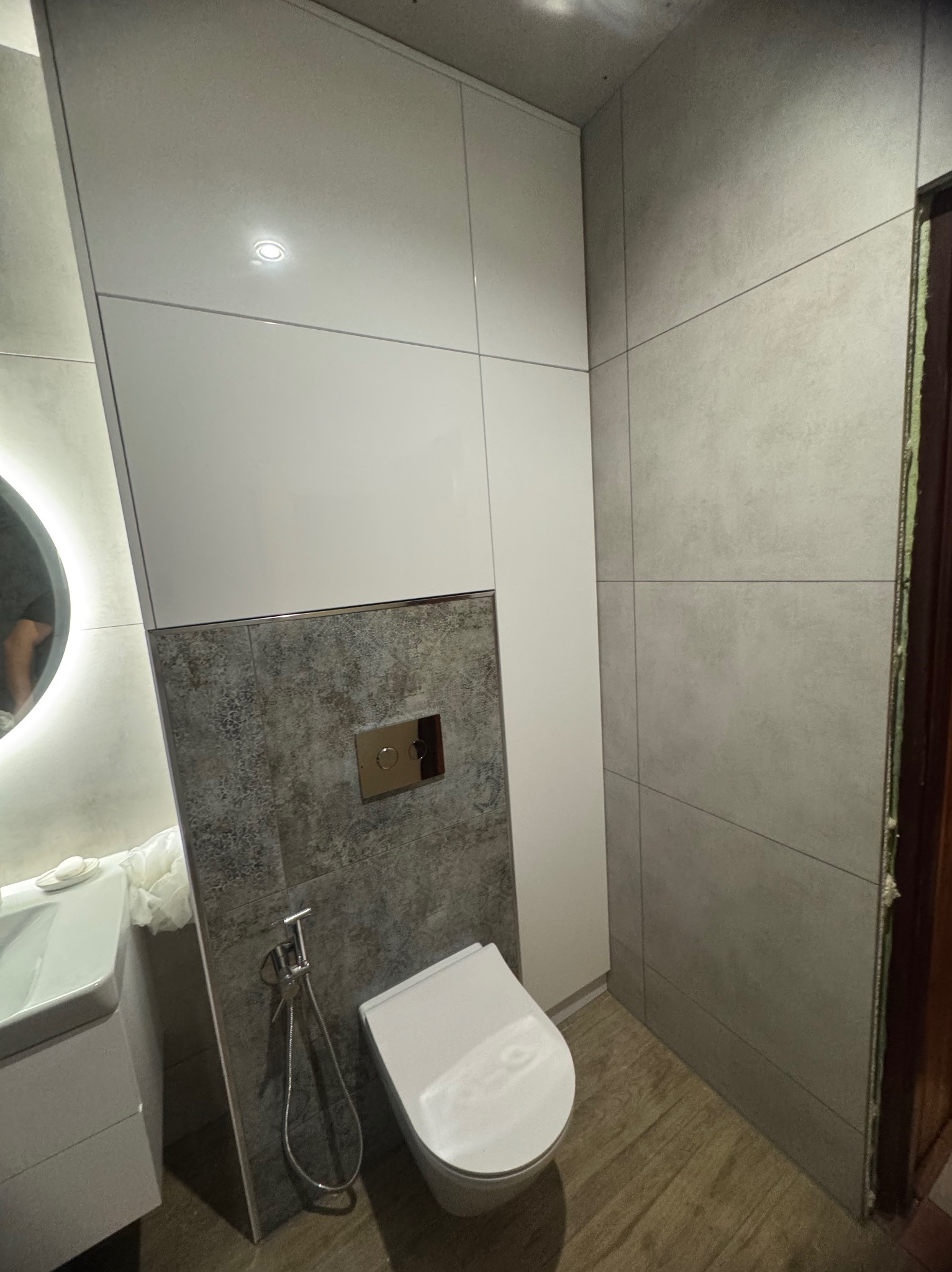 Nowoczesna łazienka z zabudową szafki nad toaletą, szara glazura, biała ceramika i okrągłe lustro z podświetleniem LED. Minimalistyczny design.