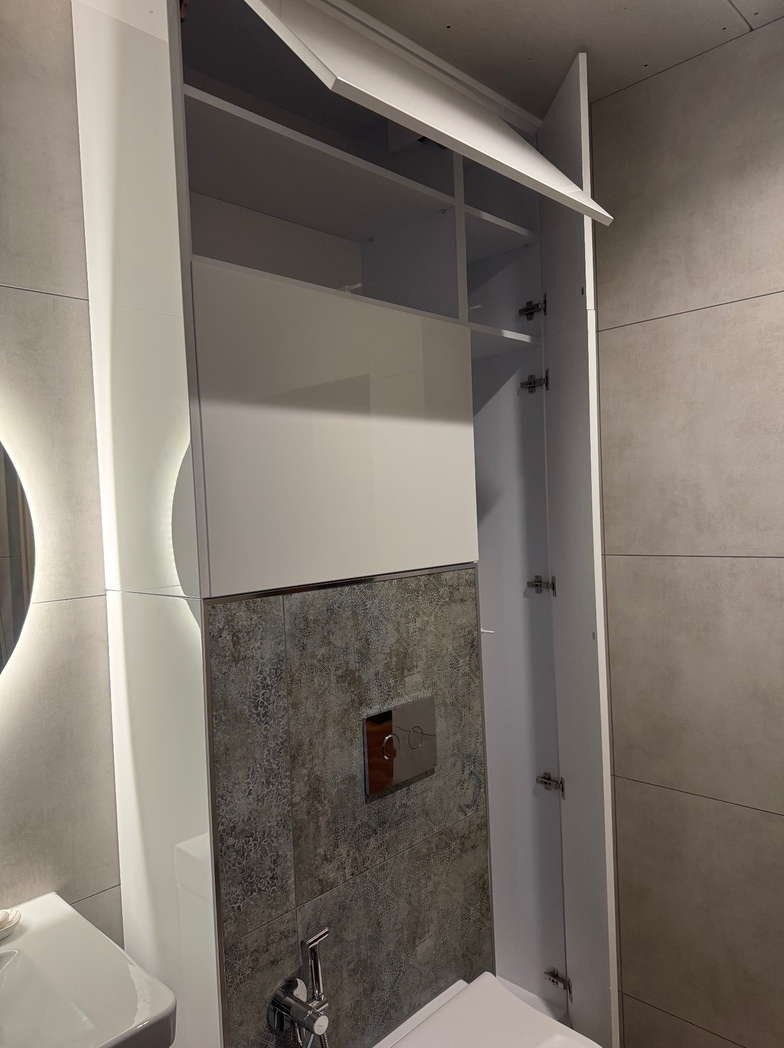 Biała szafka wnękowa w łazience z otwartymi drzwiczkami, widoczne półki. Na ścianie szare płytki z dekoracyjnym wzorem. U dołu toaleta i bidetka.