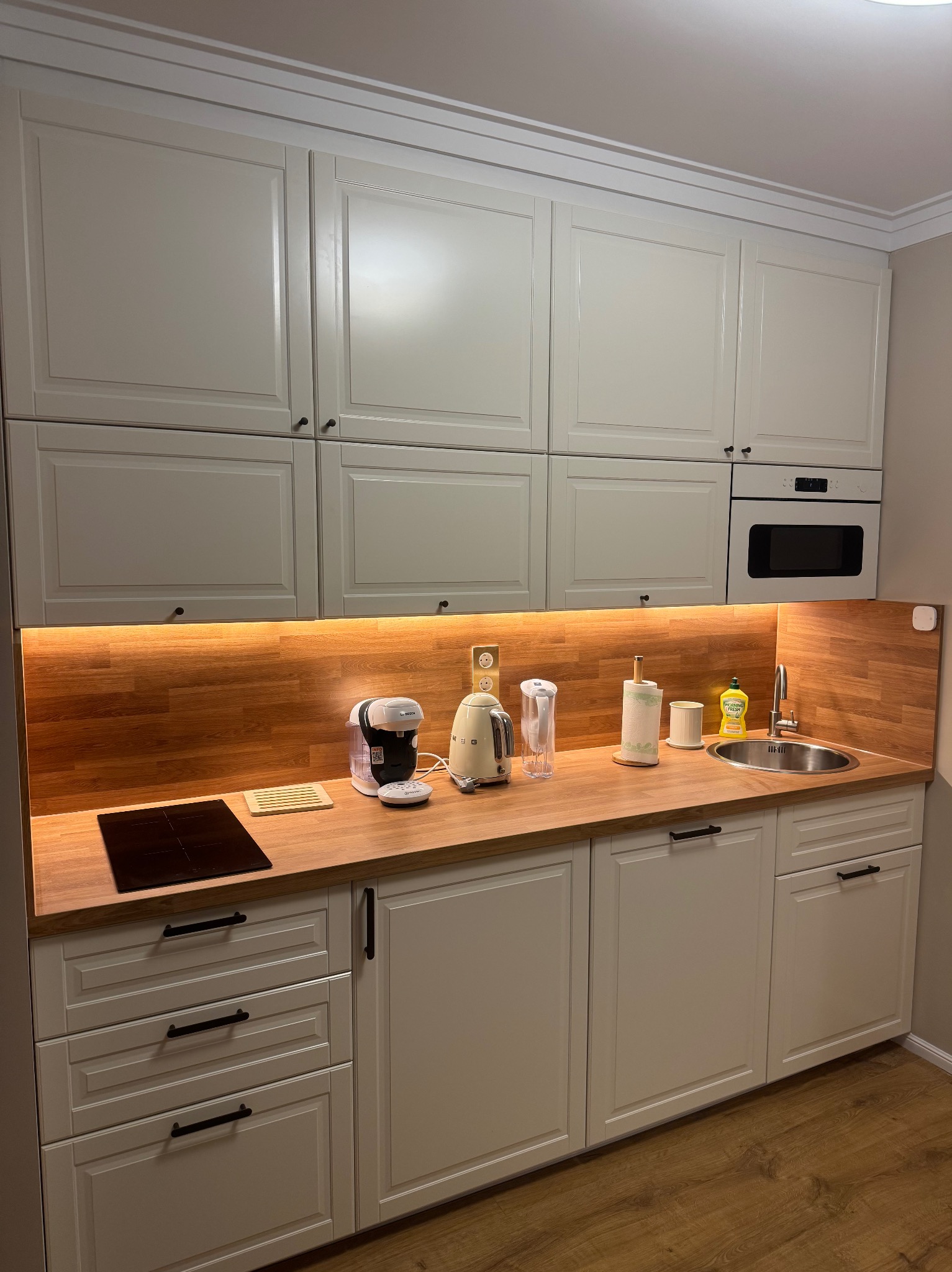 Jasna kuchnia na wymiar z drewnianym blatem i oświetleniem LED pod szafkami. Widoczny zlew, płyta indukcyjna i sprzęty AGD. Fronty szafek frezowane, uchwyty czarne.