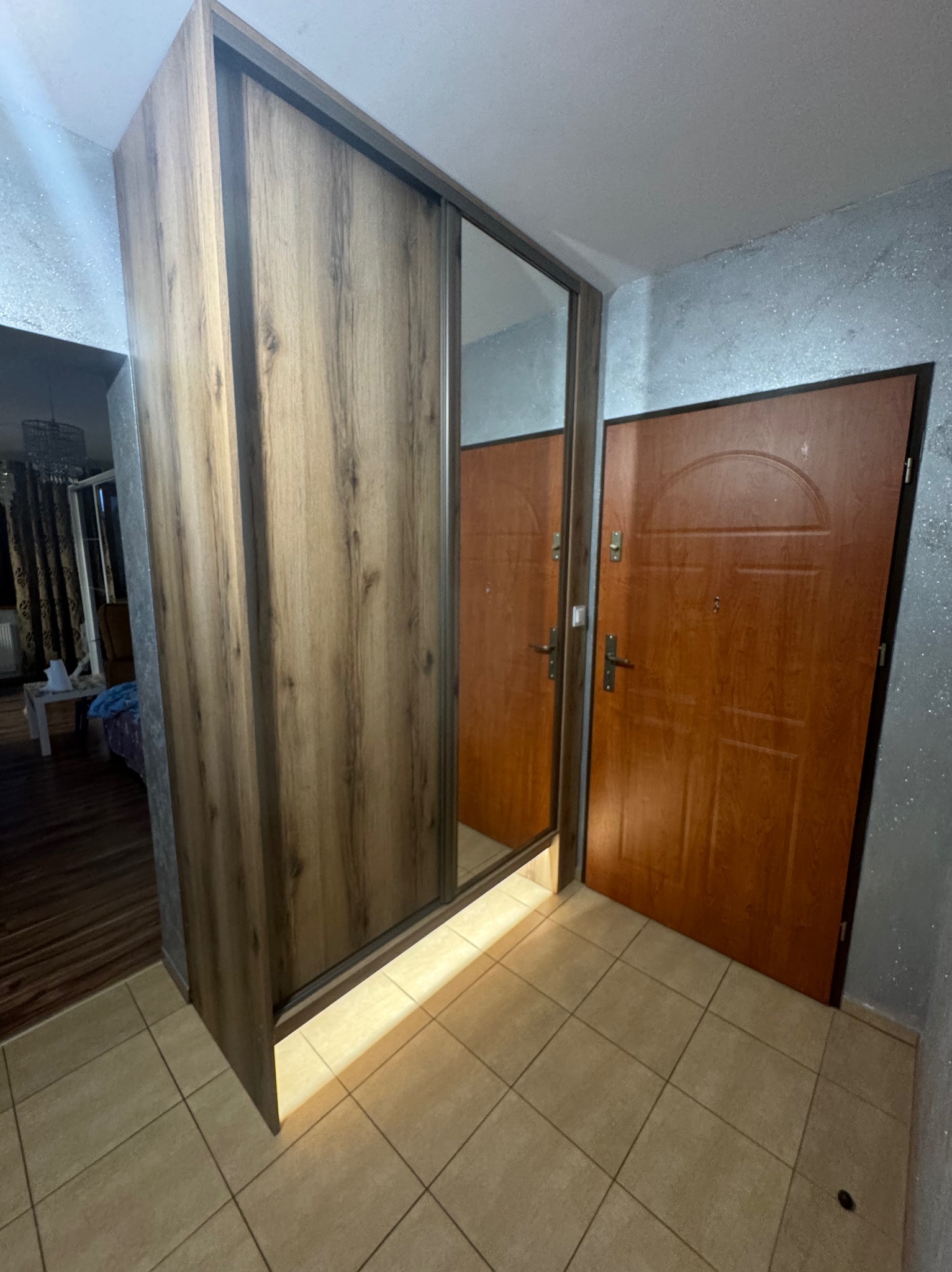Wnętrze przedpokoju z szafą wnękową z przesuwnymi drzwiami z lustrem i drewnianym wykończeniem, obok brązowe drzwi wejściowe. Widoczna podłoga z płytek.