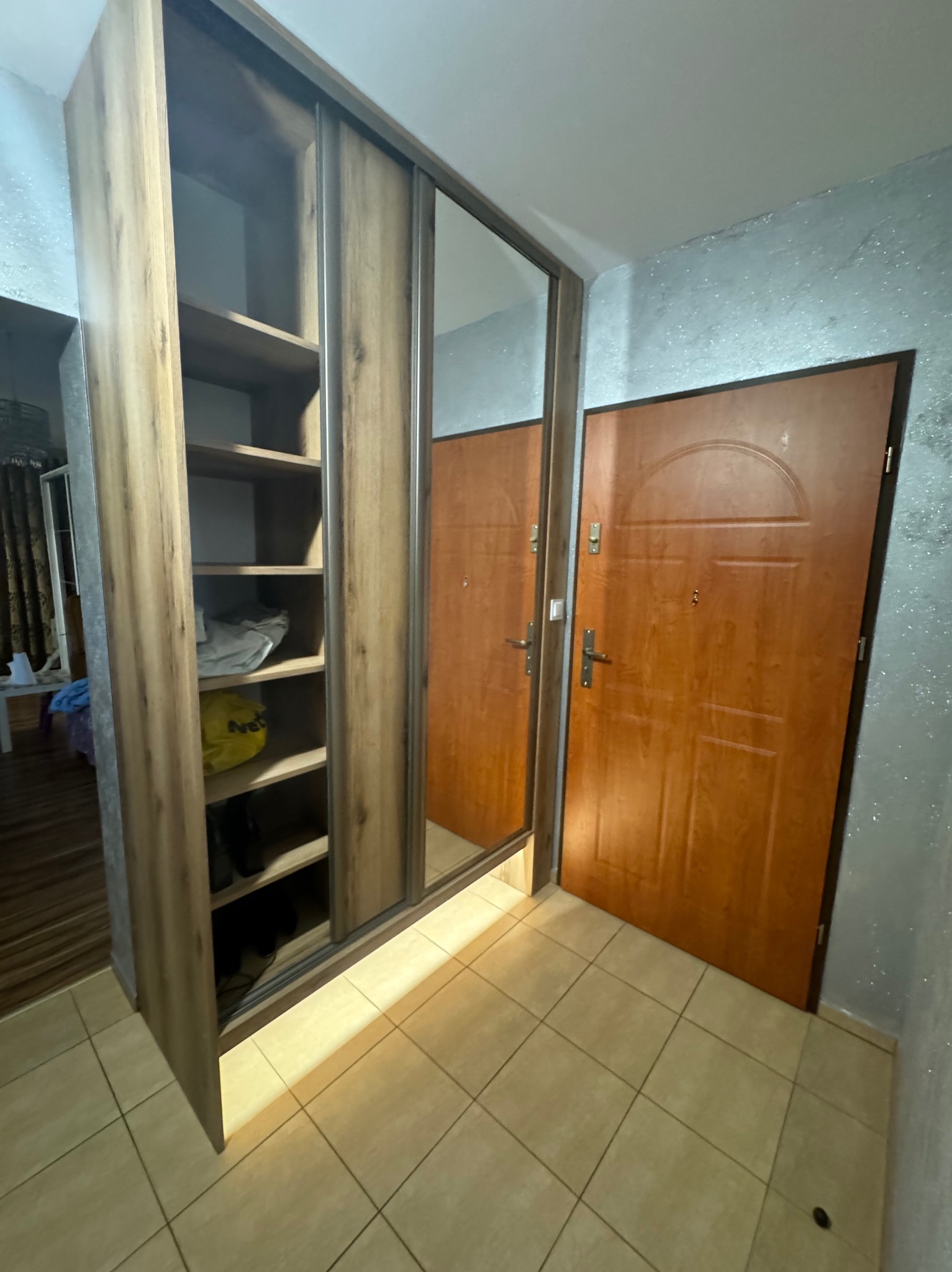 Szafa wnękowa z lustrzanymi drzwiami przesuwnymi, drewnopodobne wykończenie, widoczne półki z zawartością, obok brązowe drzwi wejściowe, płytki podłogowe.