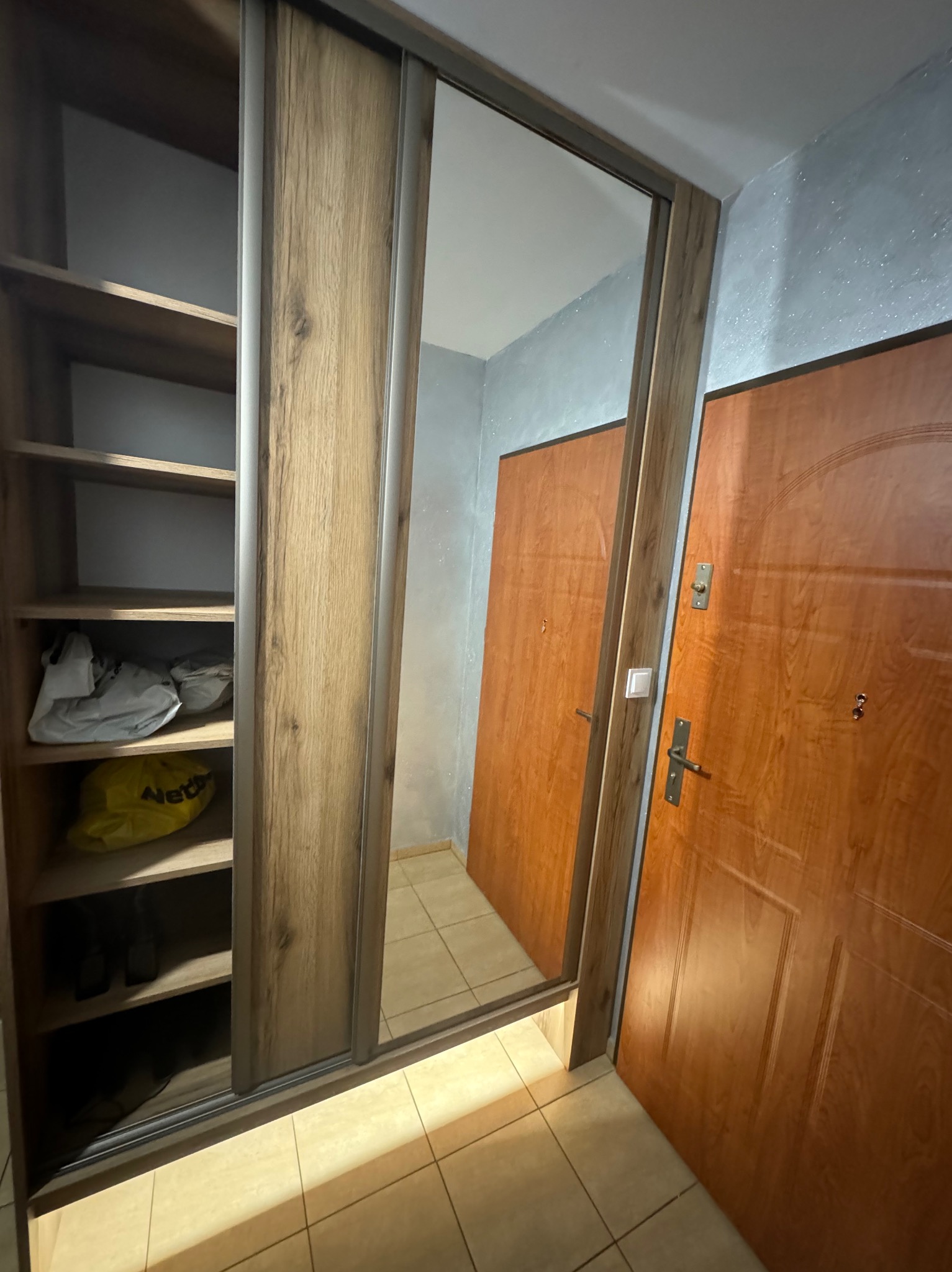 Szafa wnękowa z lustrem i drewnianymi przesuwnymi drzwiami, widok na półki z ubraniami i odbicie w lustrze, obok drewniane drzwi wejściowe.