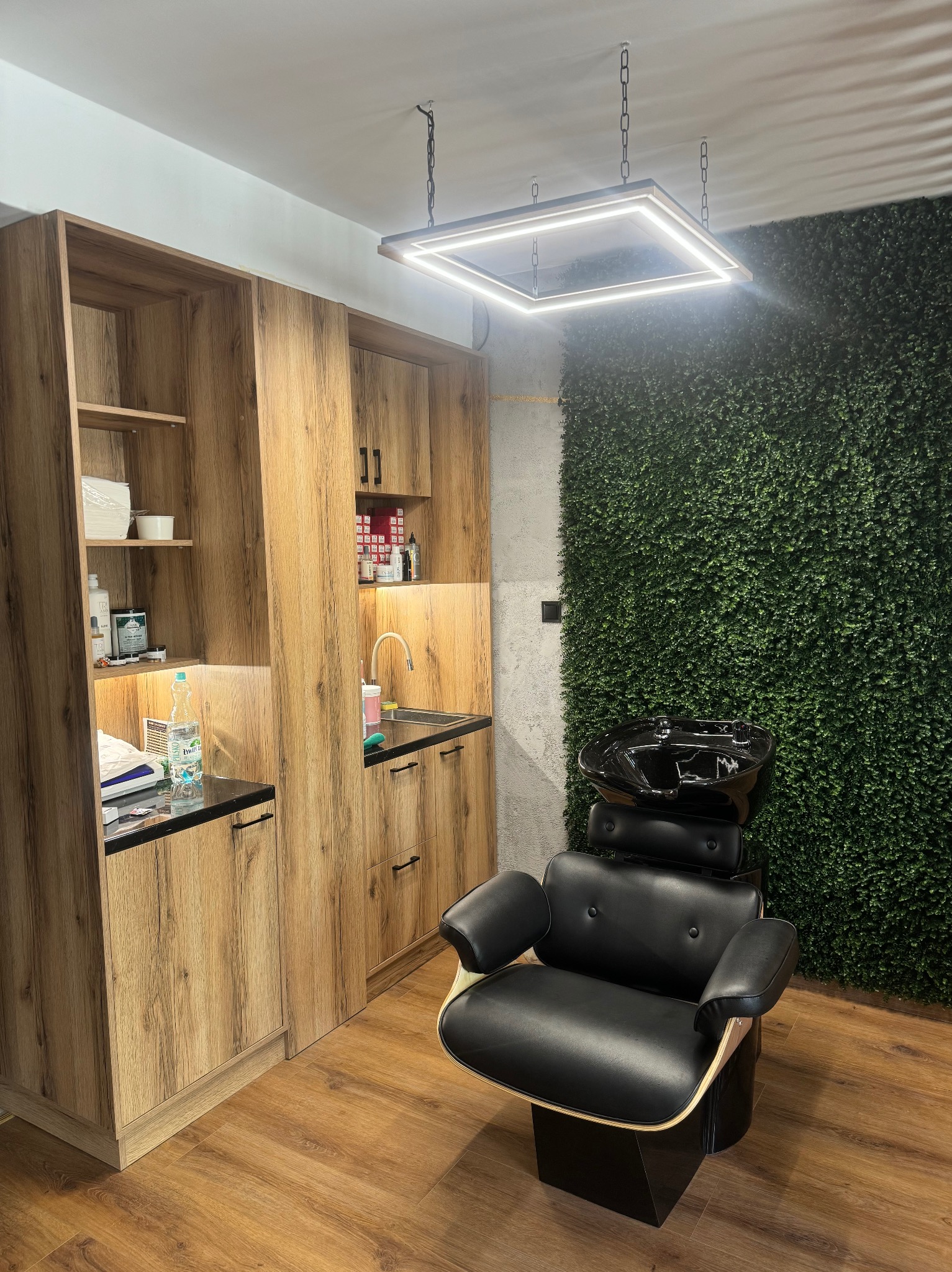 Nowoczesne meble w salonie fryzjerskim: szafki z drewnopodobnym fornirem, czarne krzesło do mycia włosów na tle ściany z zielonej roślinności.