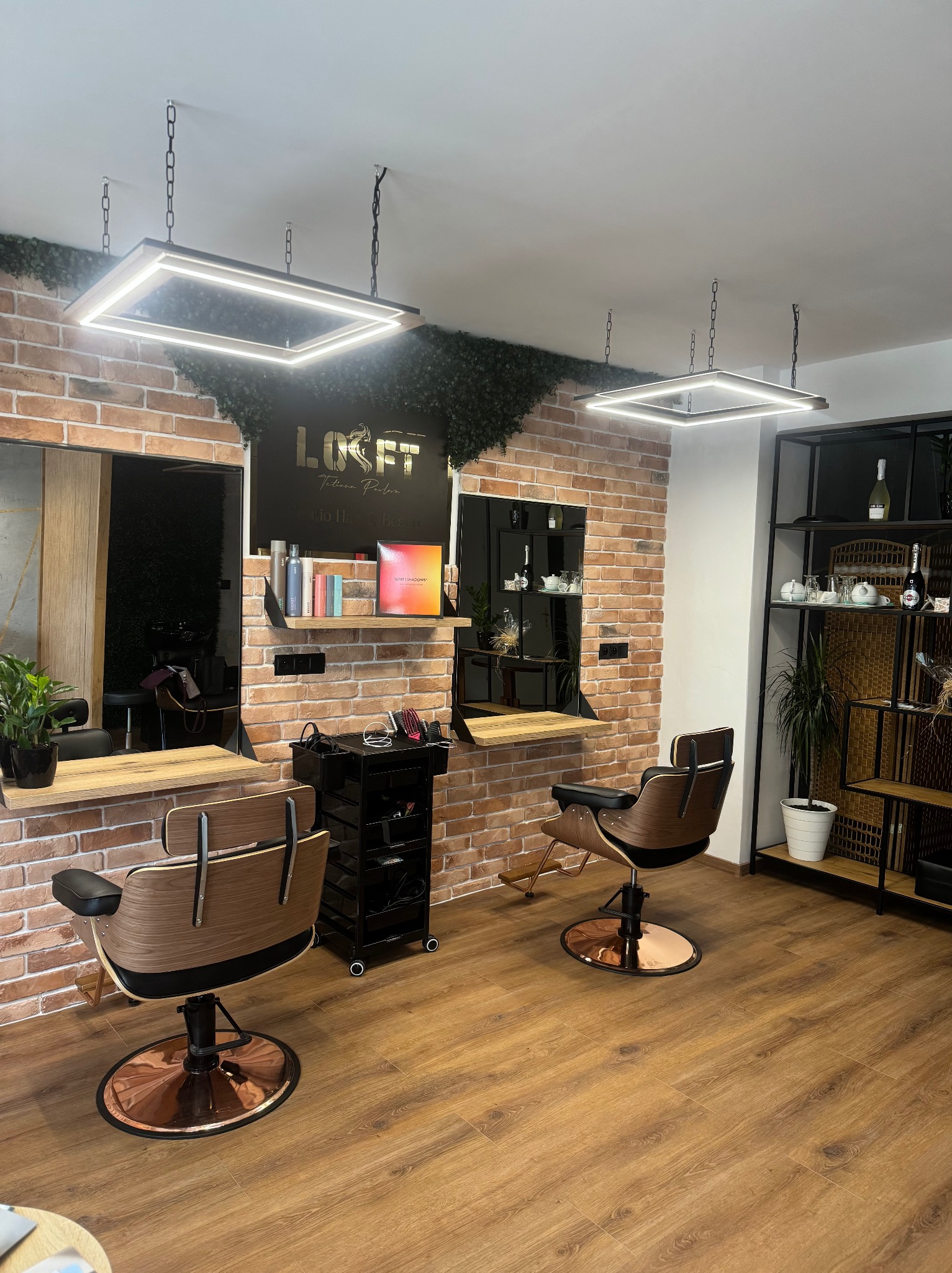 Nowoczesne wnętrze salonu fryzjerskiego z ceglaną ścianą, drewnianymi elementami i designerskimi lampami. Widoczne stanowiska pracy z lustrami i wygodnymi fotelami.