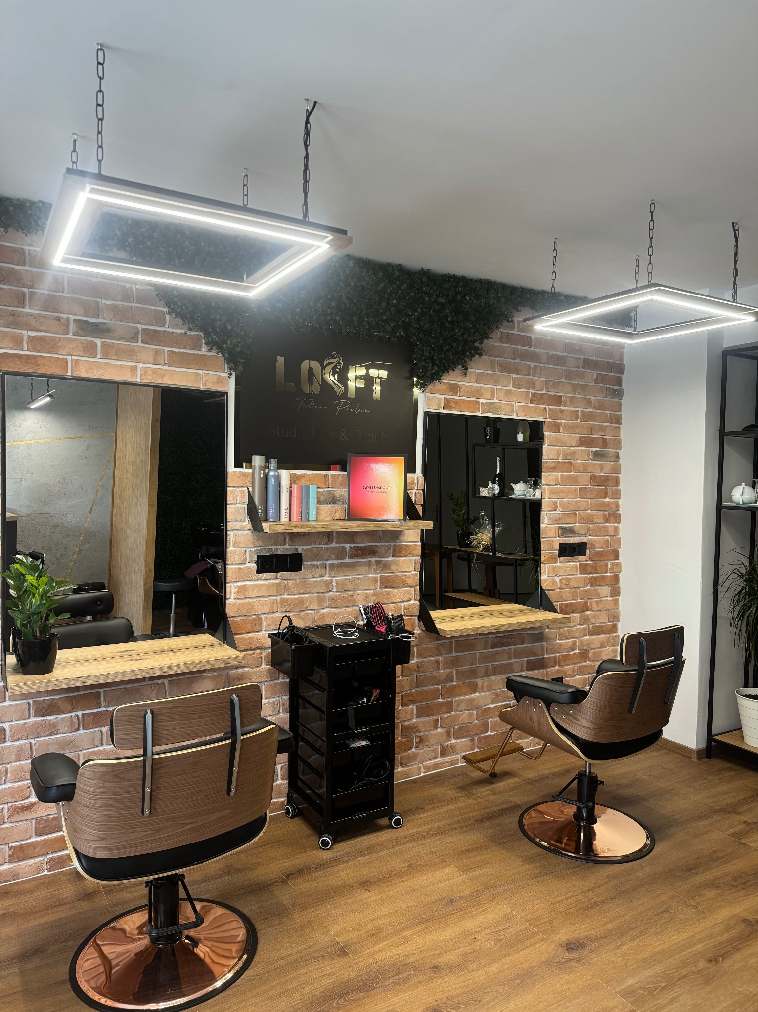 Nowoczesne stanowiska fryzjerskie z drewnianymi blatami i lustrami na tle ceglanej ściany z zielonym akcentem, oświetlone kwadratowymi lampami wiszącymi.