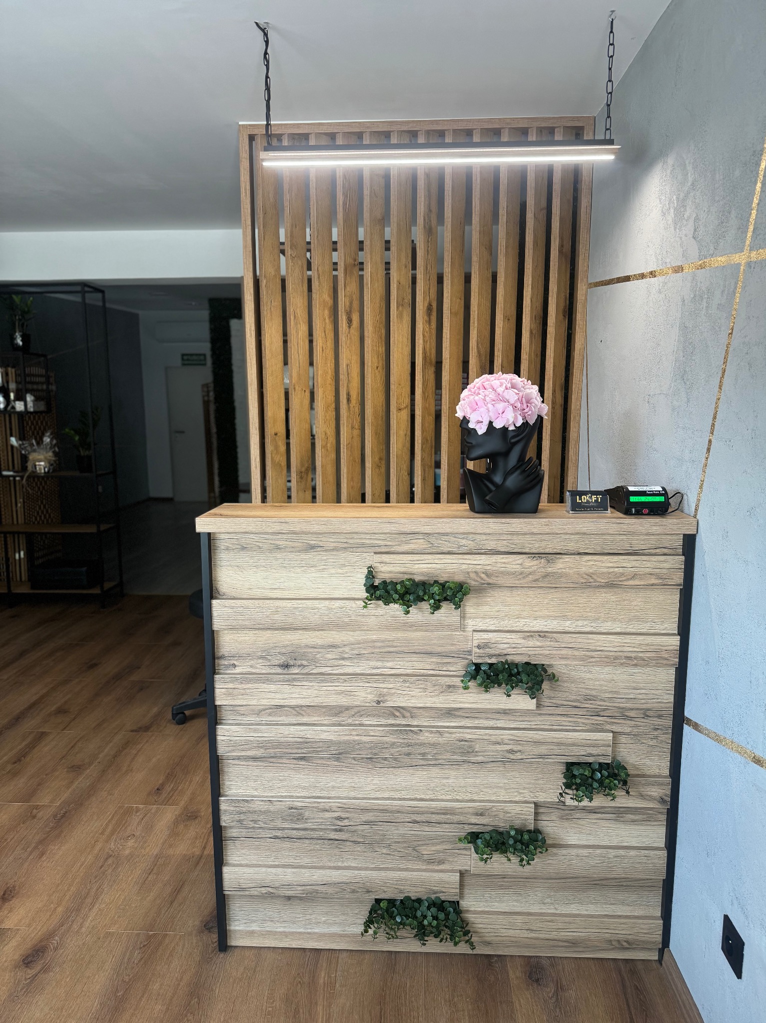 Drewniana lada recepcyjna z dekoracyjnymi roślinami i oświetleniem LED, uzupełniona o drewnianą ściankę działową. Nowoczesny design z elementami loftowymi.
