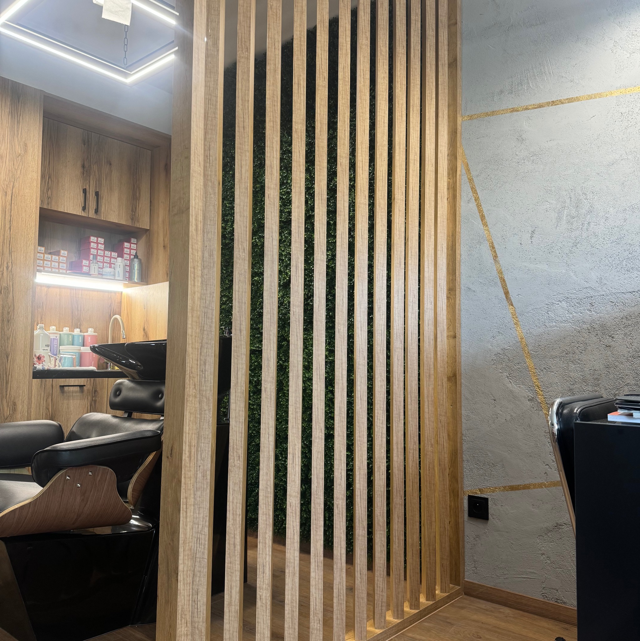 Nowoczesna drewniana ścianka działowa z pionowych listew w salonie fryzjerskim, oddzielająca strefę mycia włosów. W tle widoczna ściana z dekoracyjnymi złotymi liniami.