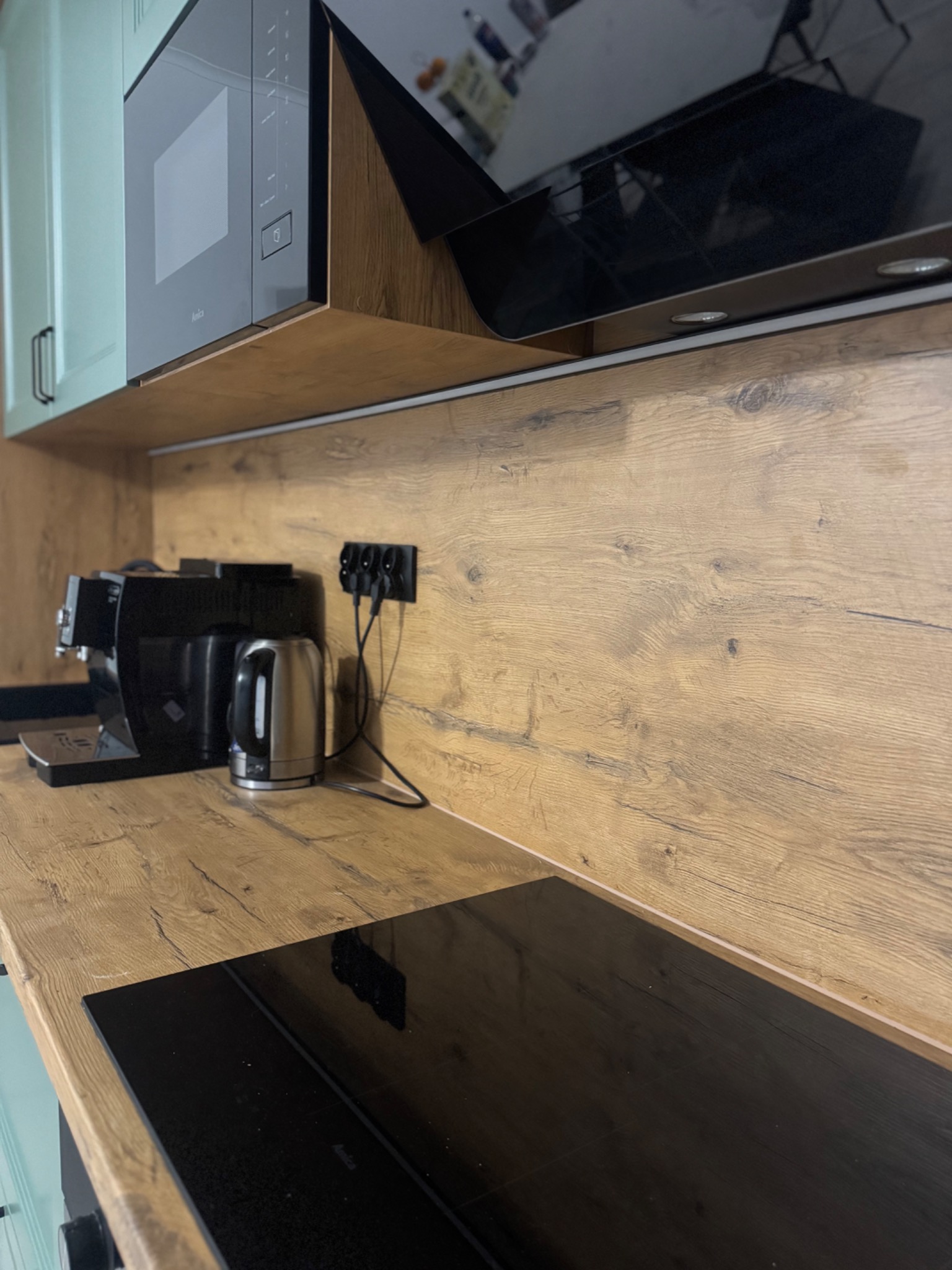 Fragment kuchni z drewnianym blatem, czarną płytą indukcyjną i sprzętami AGD. Uwagę zwraca naturalny odcień drewna i minimalistyczny design.