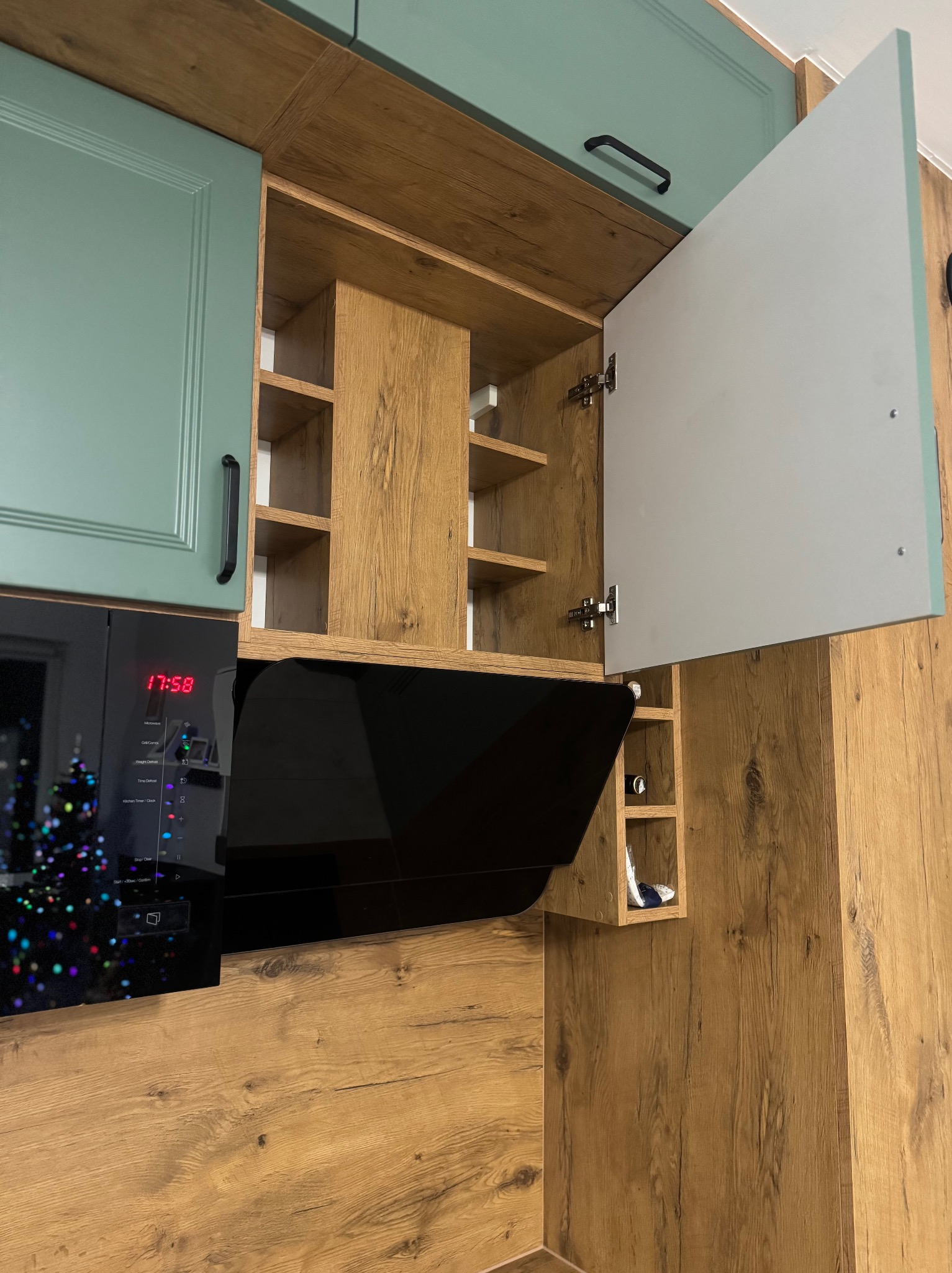 Nowoczesna kuchnia z drewnianymi szafkami, zielonymi frontami i otwartą szafką z półkami. Widoczny czarny okap i mikrofalówka z wyświetlaczem czasu.