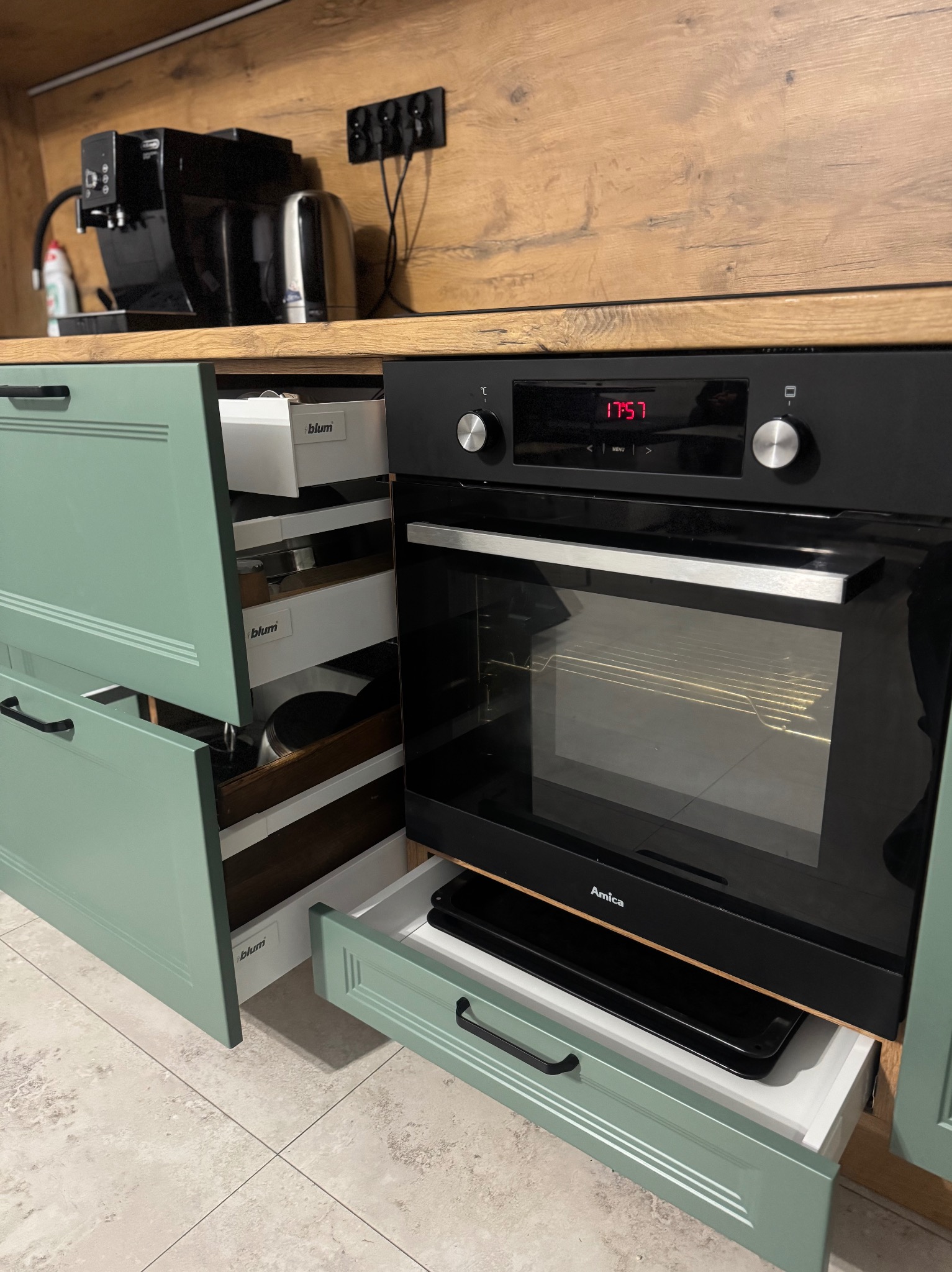 Szczegółowe ujęcie zabudowy kuchennej na wymiar: pastelowe, frezowane fronty szuflad Blum, piekarnik Amica i drewniany blat. Widoczne wyposażenie szuflad.