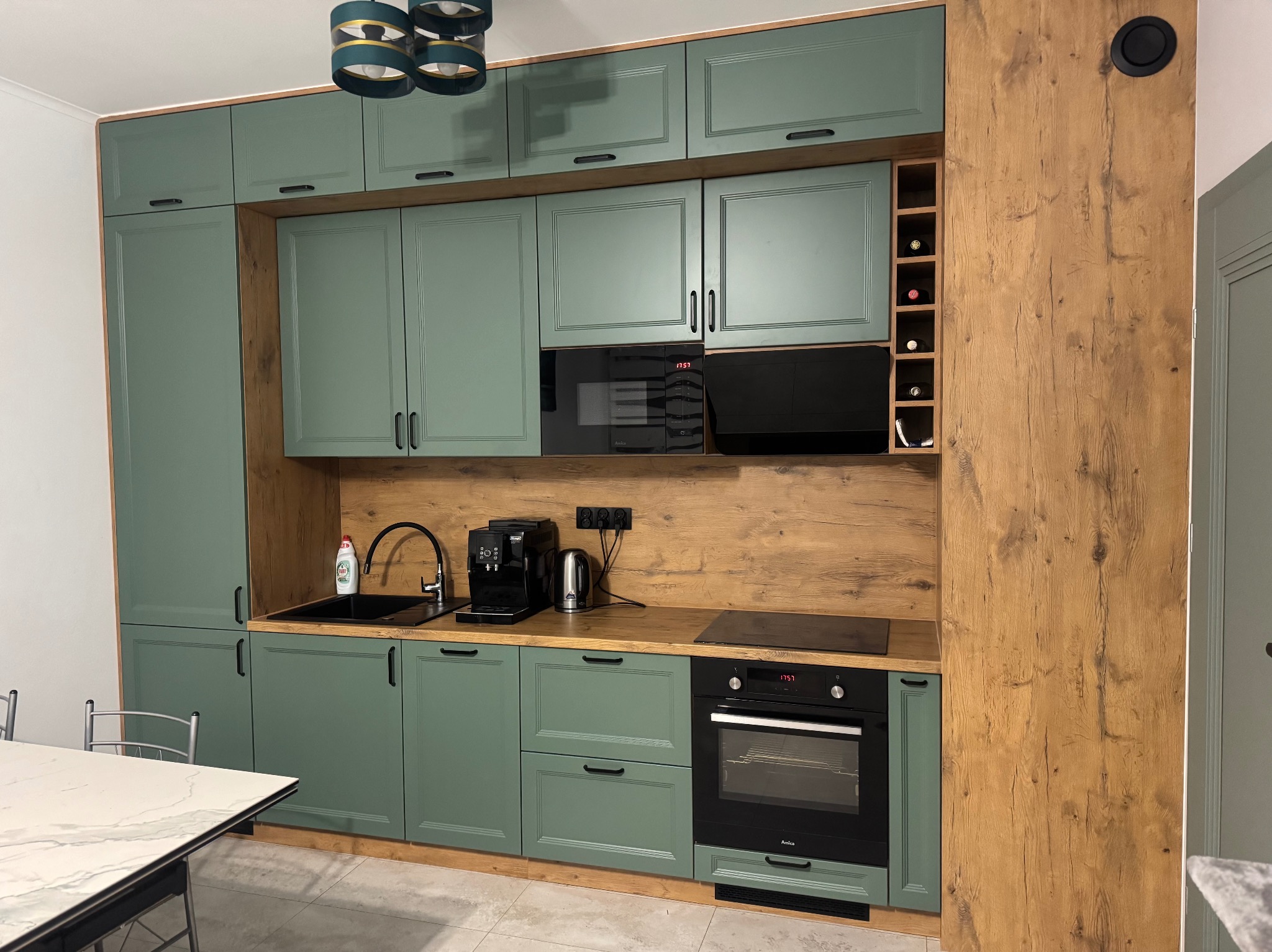 Nowoczesna kuchnia z zielonymi frontami i drewnianym blatem. Widoczny piekarnik, płyta indukcyjna, ekspres do kawy i wbudowana szafka na wino. Całość oświetlona designerską lampą.