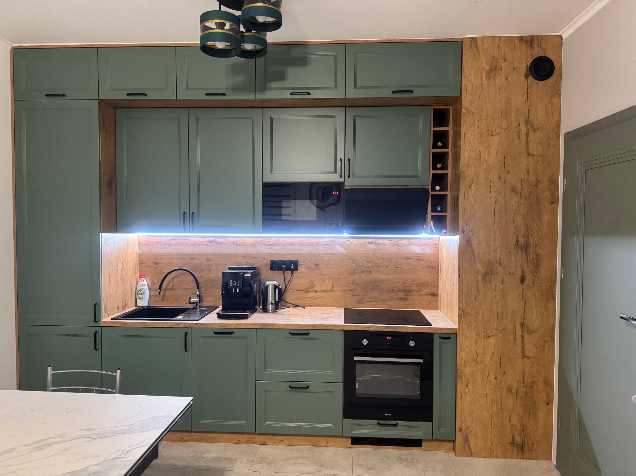 Nowoczesna kuchnia w odcieniach zieleni i drewna z czarnymi akcentami, oświetleniem LED podszafkowym i eleganckim wykończeniem, z widocznym ekspresem do kawy.