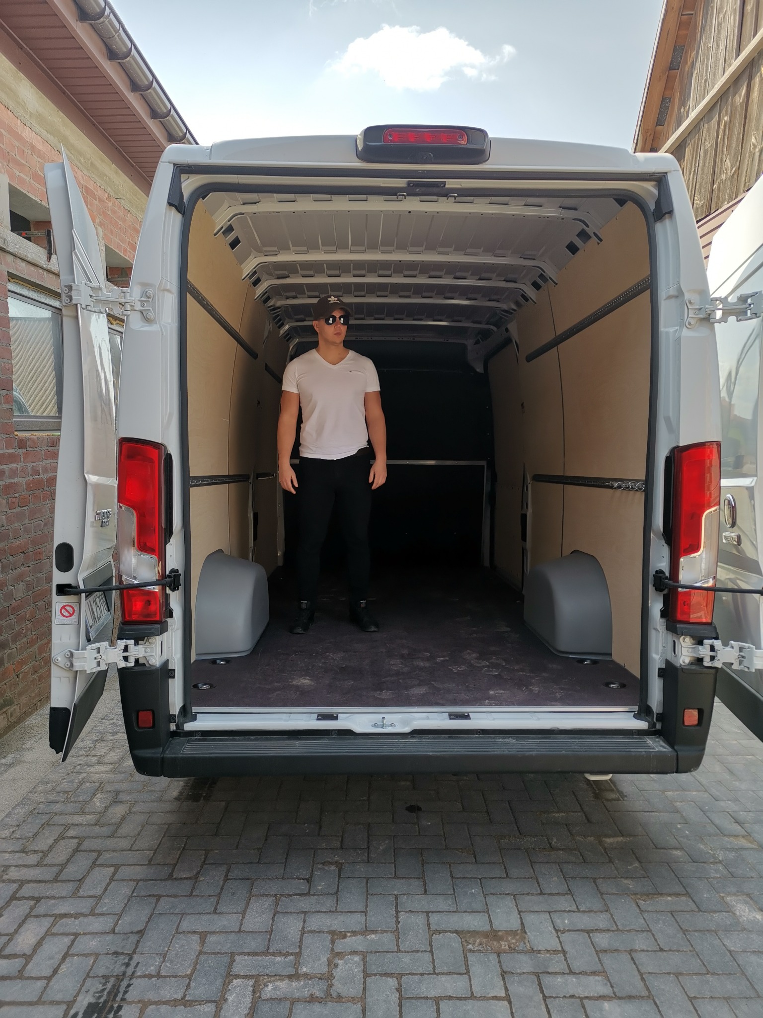 Mężczyzna w okularach słonecznych i czarnych spodniach stoi w pustej przestrzeni ładunkowej białego dostawczego busa z otwartymi tylnymi drzwiami, widok z tyłu na kostce brukowej.