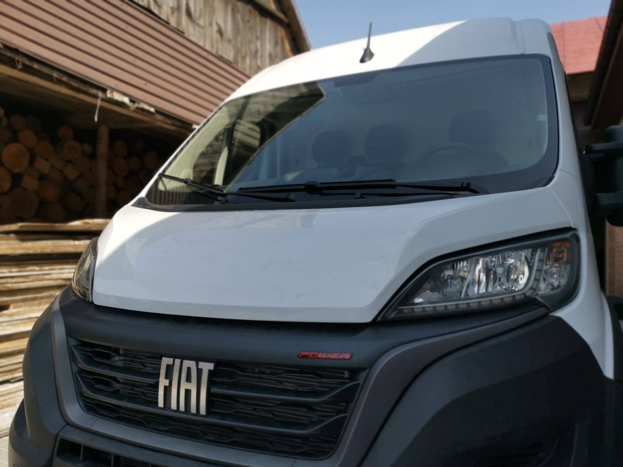 Przód białego busa marki Fiat z napisem POWER na tle drewnianego budynku i ułożonego drewna.