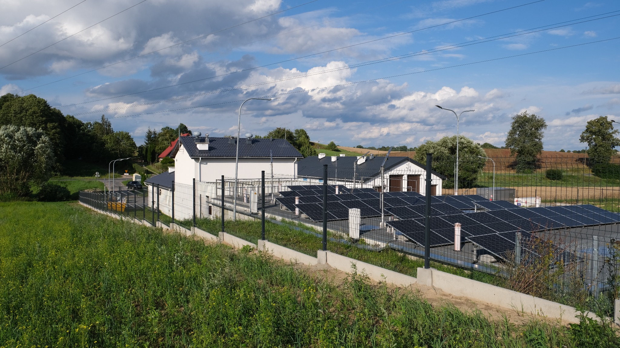 Kompleks budynków z panelami słonecznymi na dachach, otoczony ogrodzeniem z siatki na betonowej podmurówce, widok z zarośniętego terenu w słoneczny dzień.