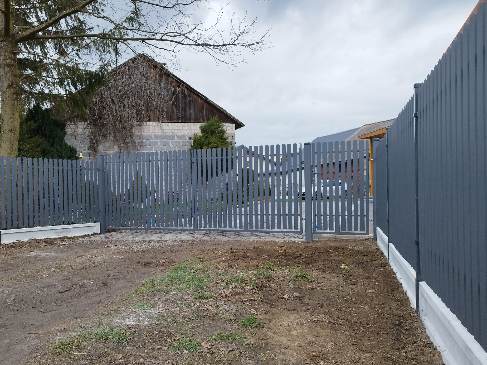 Szary, metalowy płot panelowy z furtką i podmurówką betonową na nierównym, trawiasto-ziemnym terenie, w tle budynek gospodarczy z cegły i drewnianym dachem.